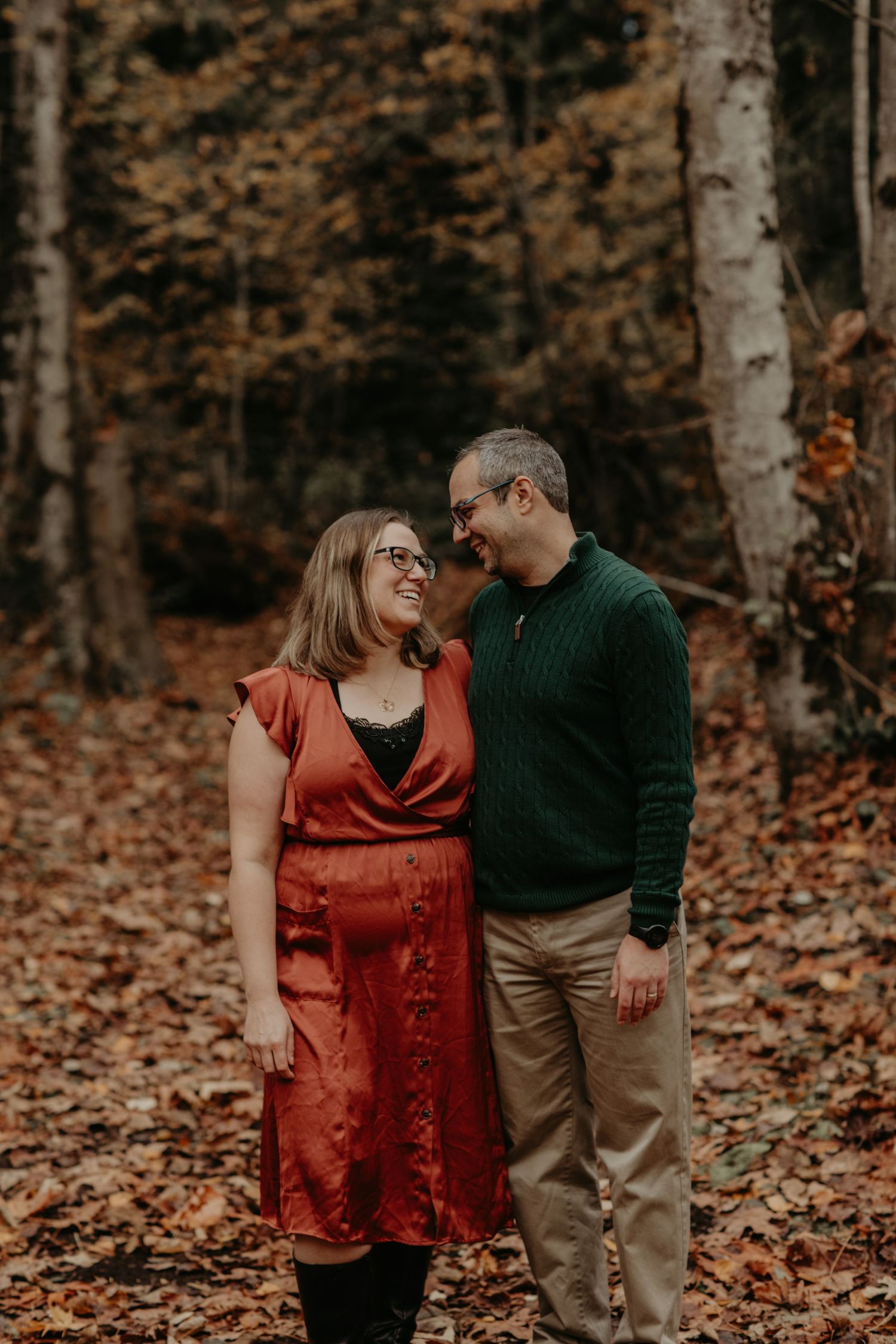 Romantic couple laughing together in autumn forest with fallen leaves, woman in red dress and man in green sweater.