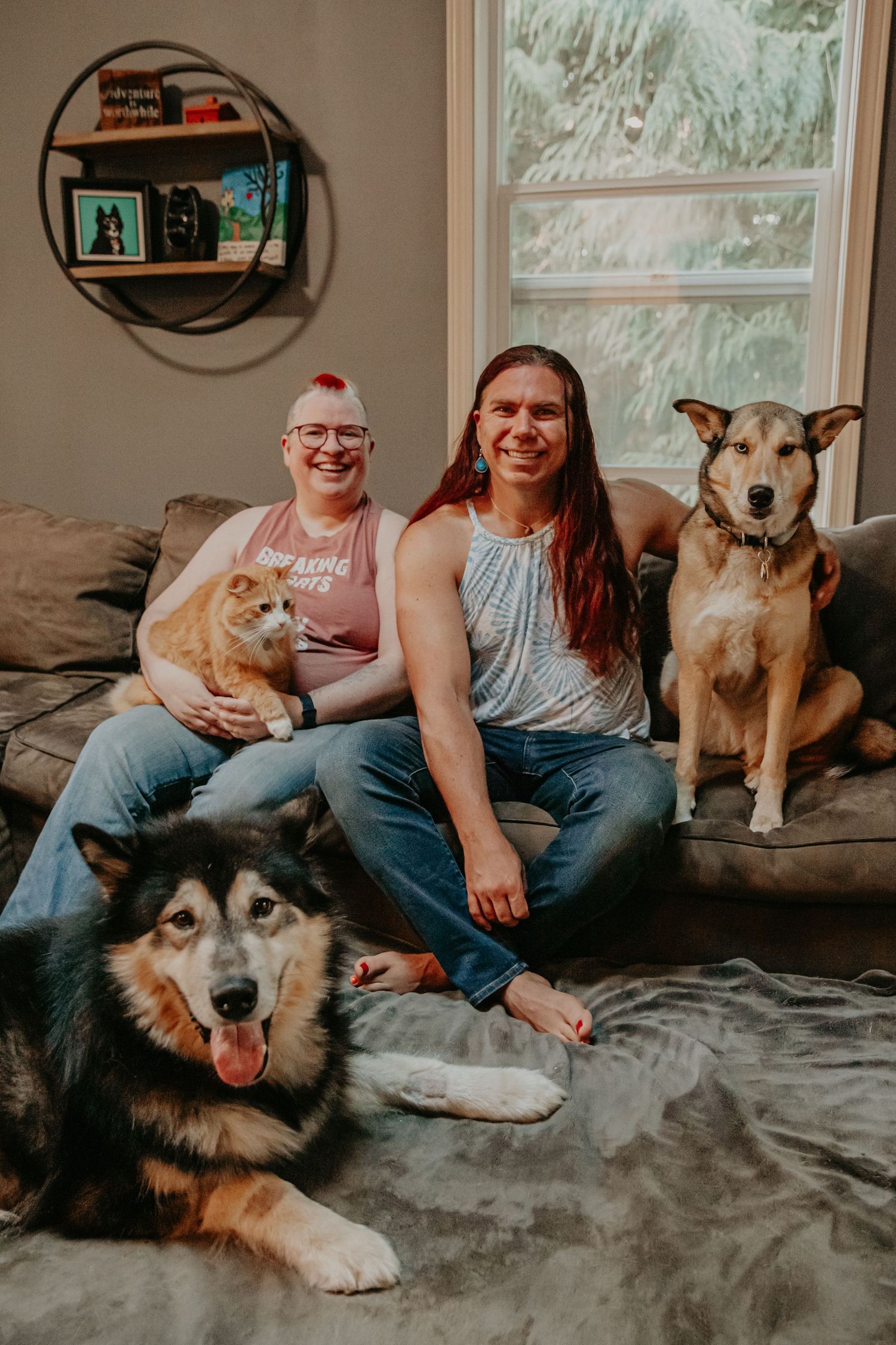 A cozy family photo on a grey couch with three pets including a ginger cat and two dogs, one being a husky.