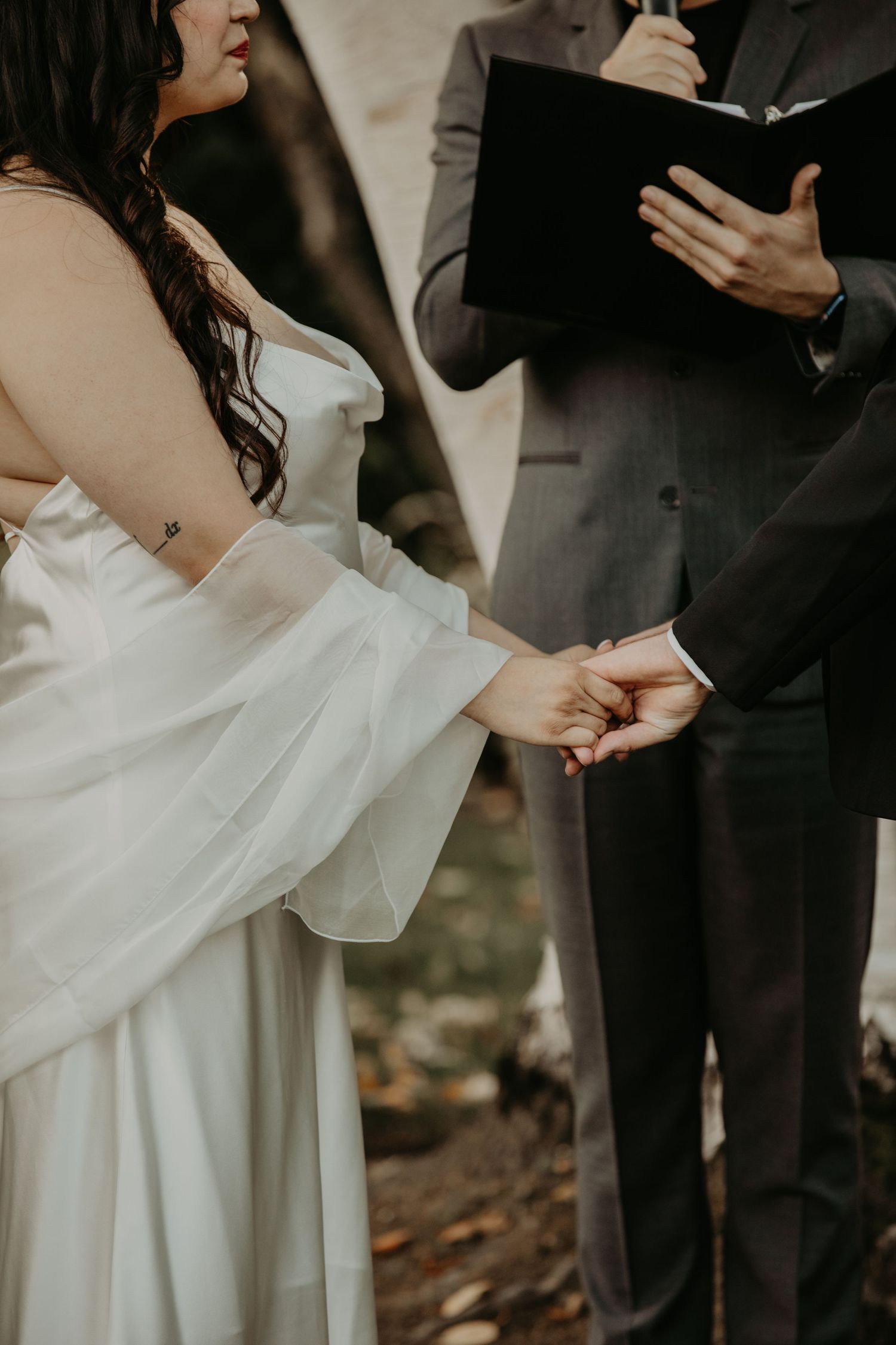 A close-up of a couple holding hands during an outdoor wedding ceremony while an officiant holds a black book.