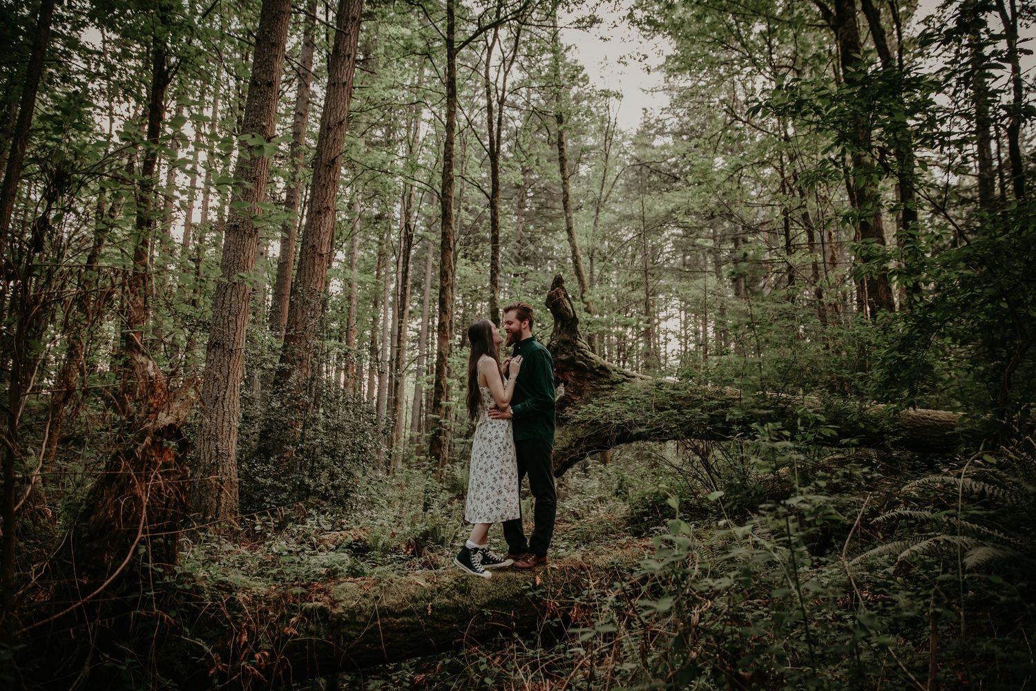 A couple stands together on a mossy path in a misty evergreen forest during an outdoor photography session.