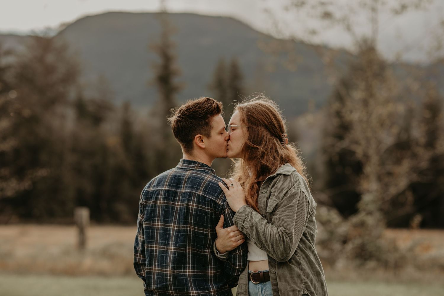 Two people in casual fall clothing embrace in a tender moment against a mountain landscape backdrop.