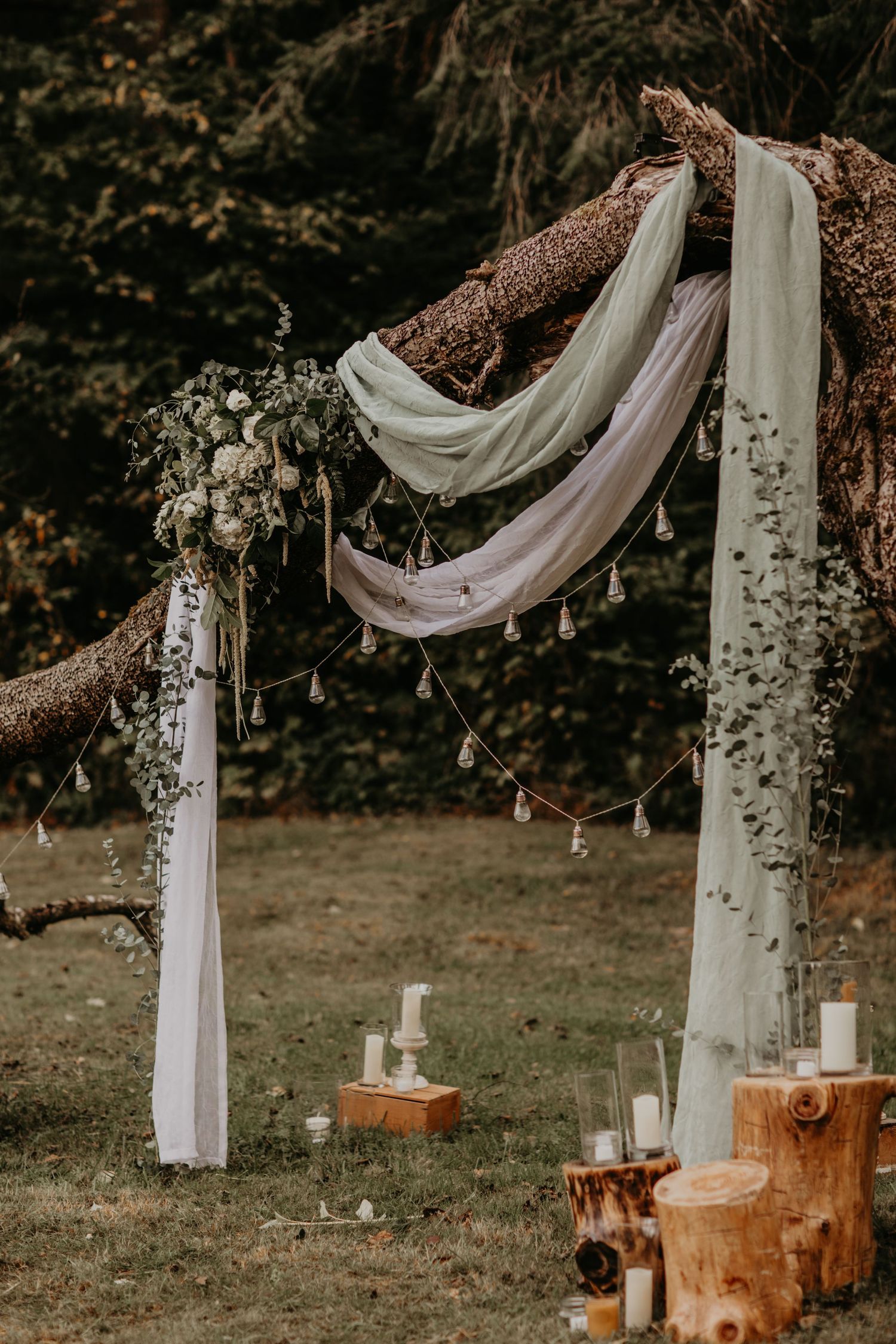 Rustic outdoor wedding ceremony setup with white draping fabric and wooden posts decorated with delicate string lights.