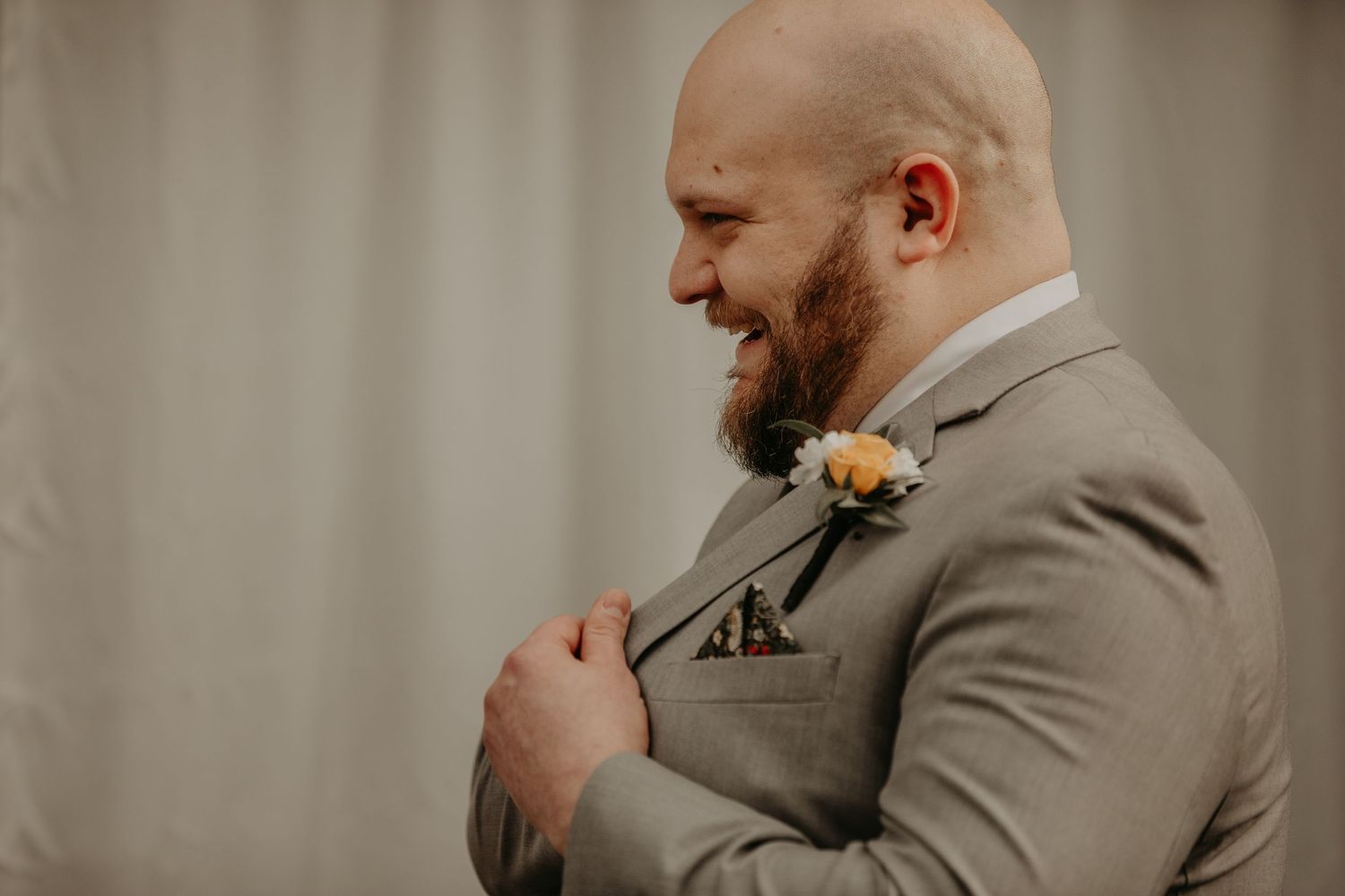 A groom in a gray suit smiles while adjusting his boutonniere before the wedding ceremony.