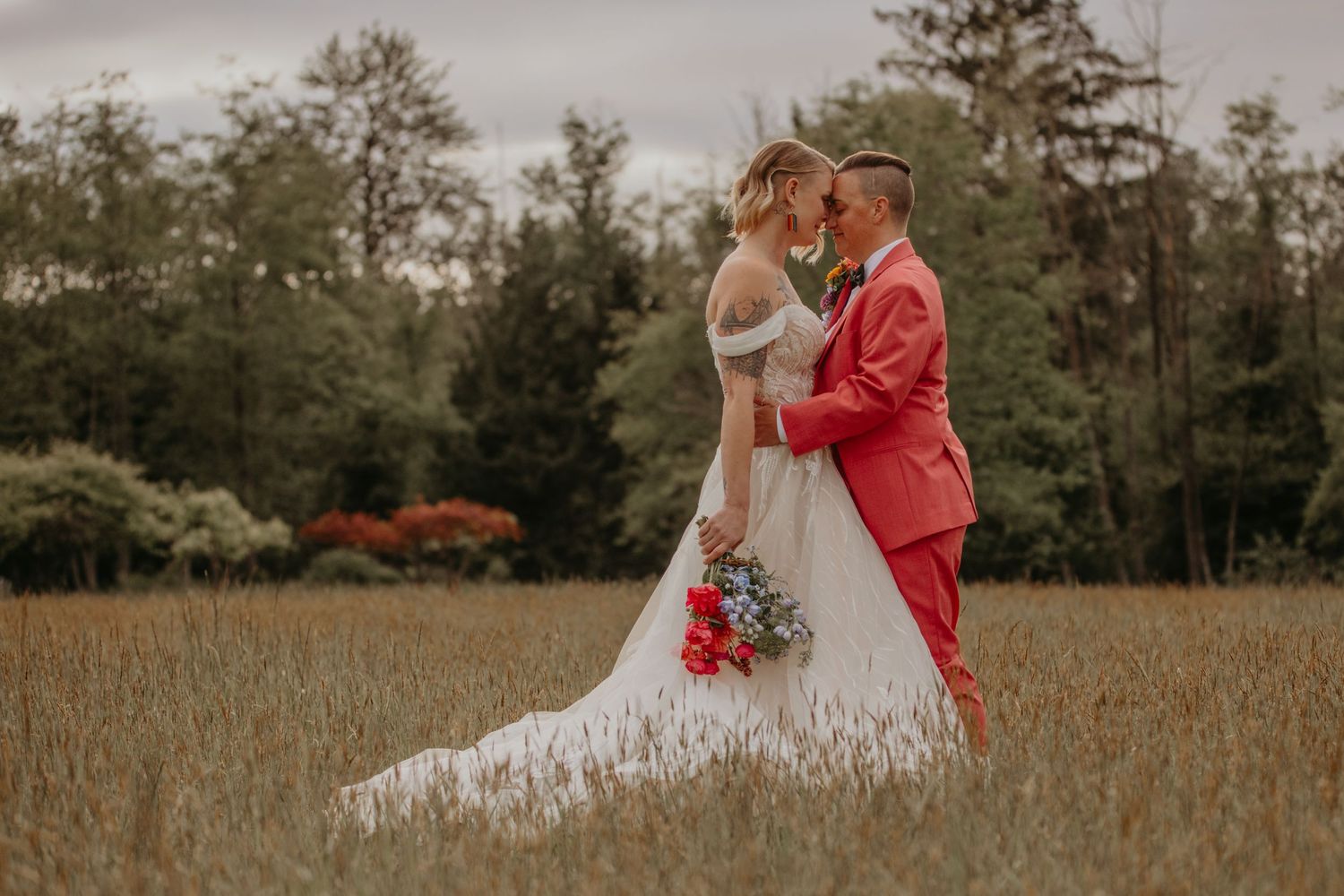 A couple embraces in a rustic field with pine trees, the person in red jacket kissing their partner in a white wedding gown.