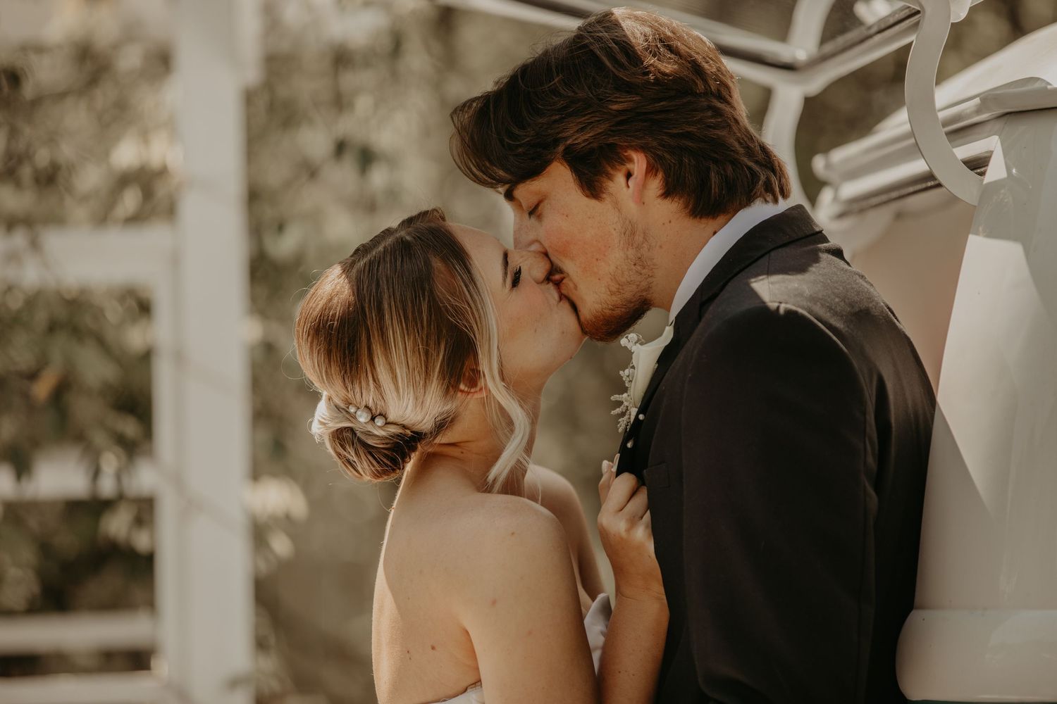 A romantic moment between newlyweds sharing a kiss under a white pergola in warm sunlight.