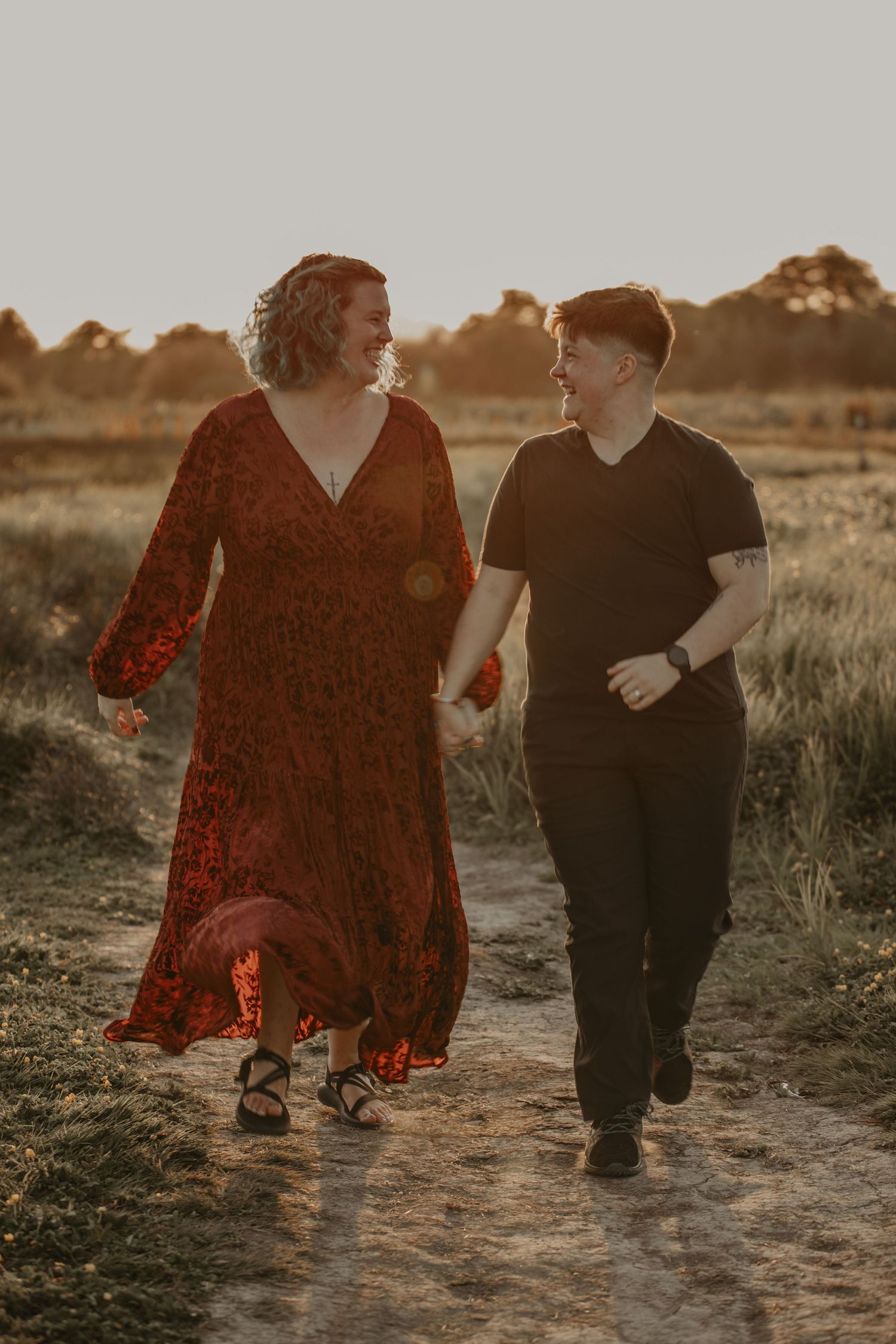 A couple holding hands walks together on a dirt path at sunset in a field during summer.