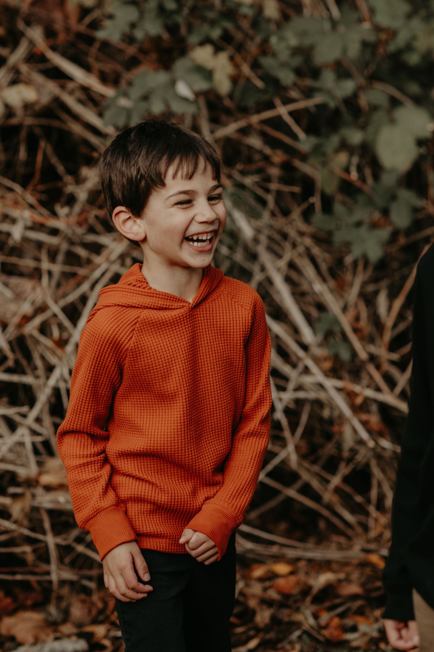 A young child wearing an orange knit sweater laughs joyfully against an autumn nature backdrop.