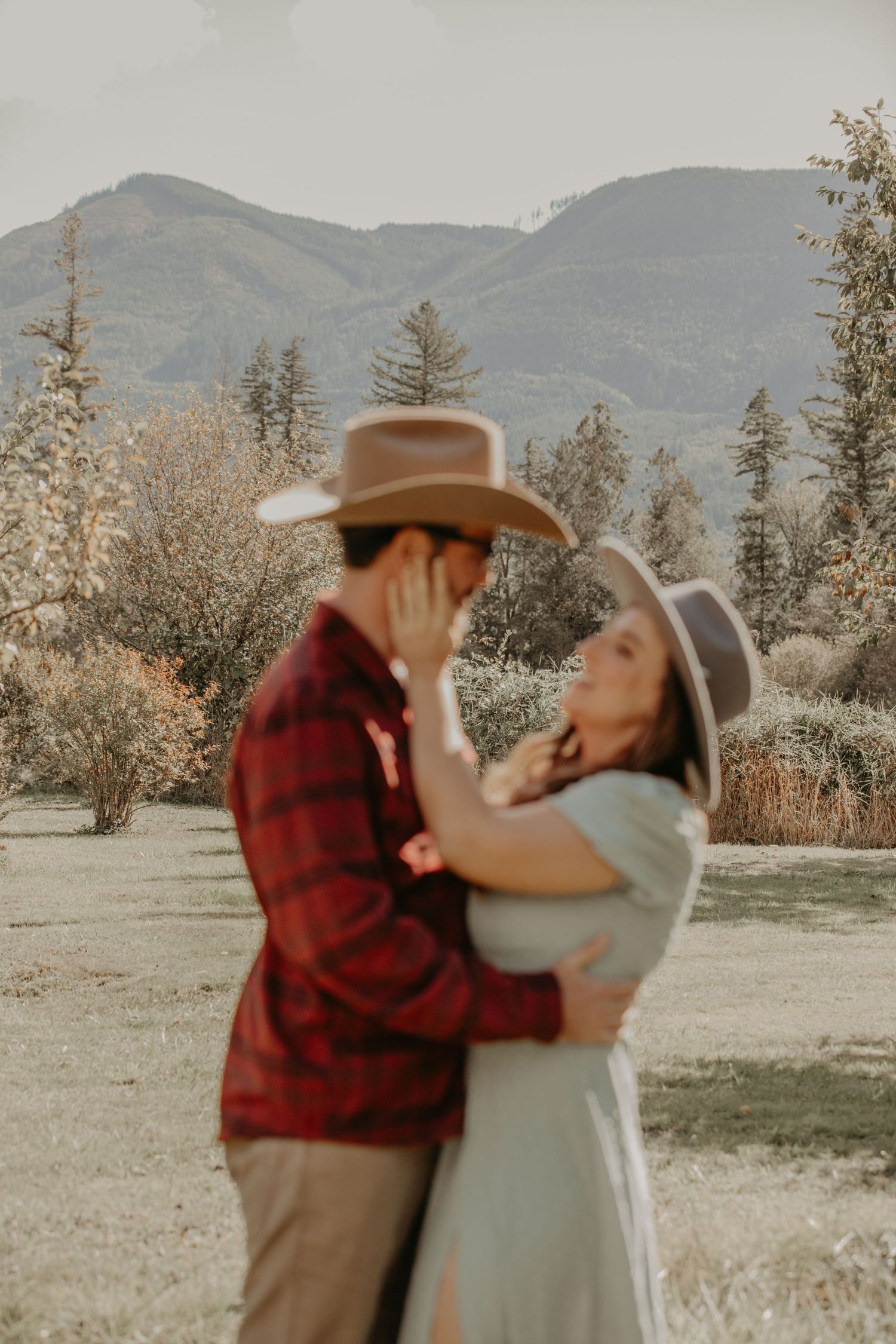 Couple in western attire embrace in an outdoor mountain setting during sunset with trees in background.