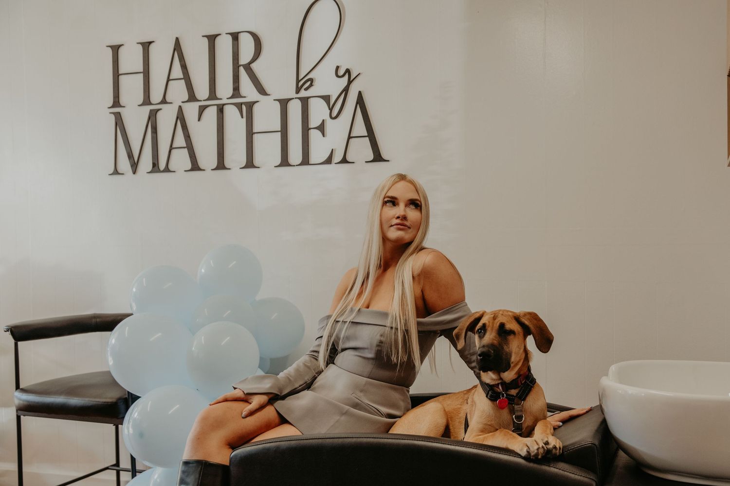 Stylish hair salon interior with blue balloons and a brown dog sitting on black bench next to salon equipment.