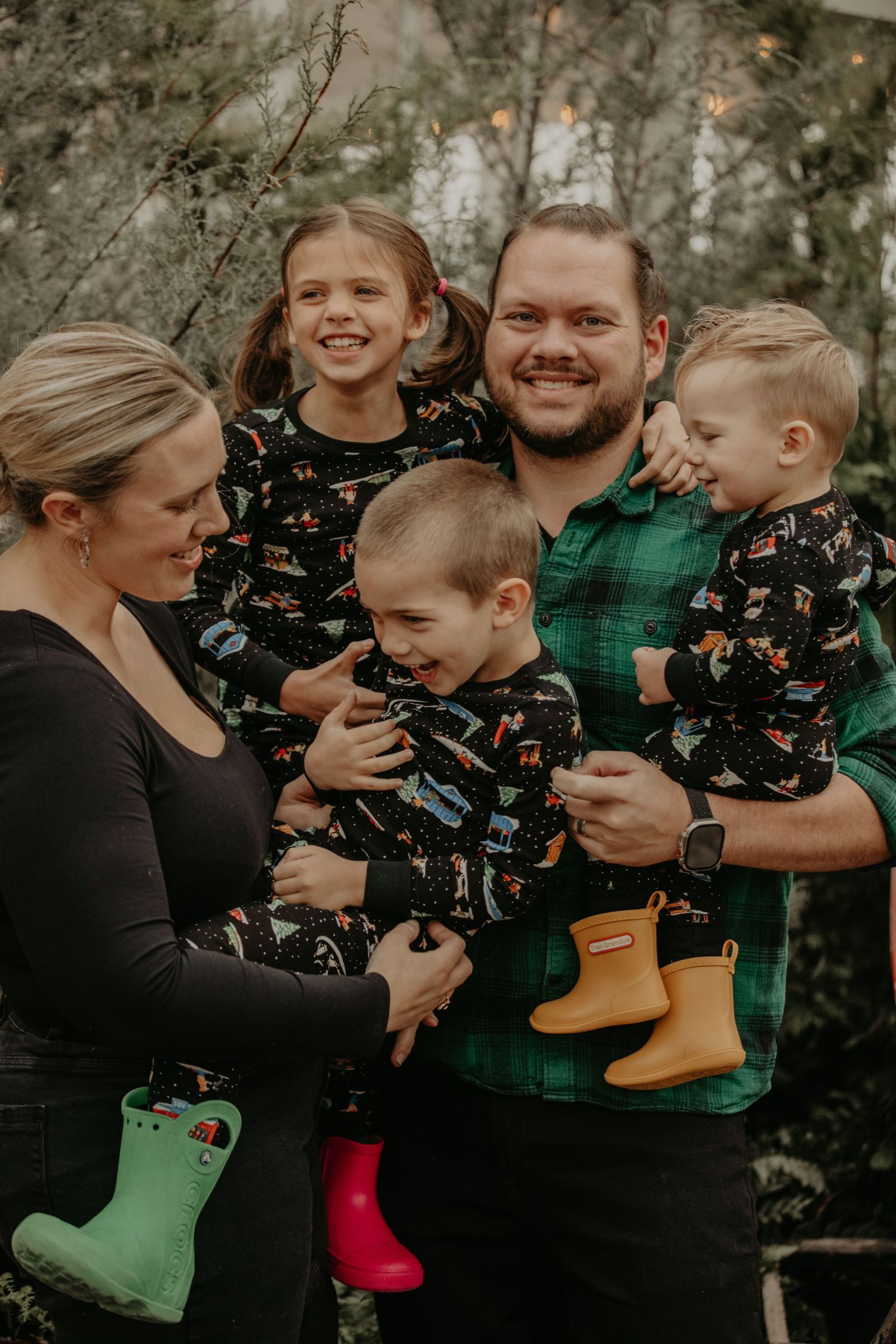 Family wearing green and black casual clothing poses together while holding colorful rain boots in a forest setting.