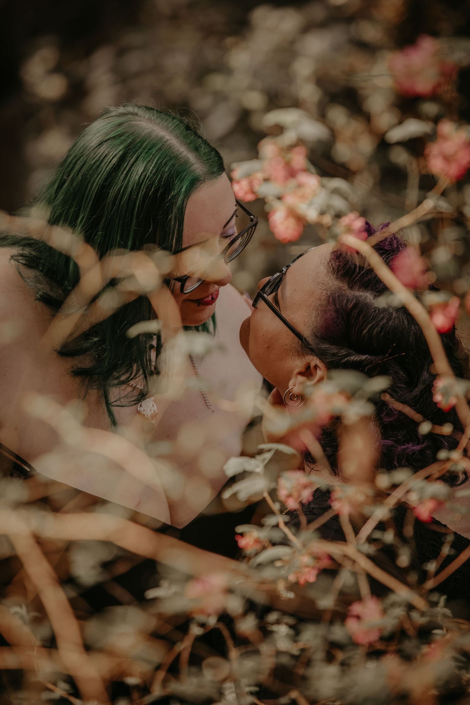 A romantic couple shares an intimate moment among blooming pink flowers in a dreamy, soft-focus photograph.