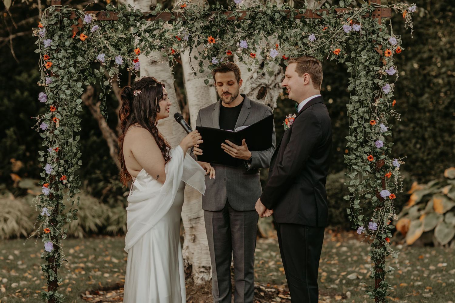 A wedding ceremony takes place under a floral arch decorated with autumn flowers and greenery outdoors.