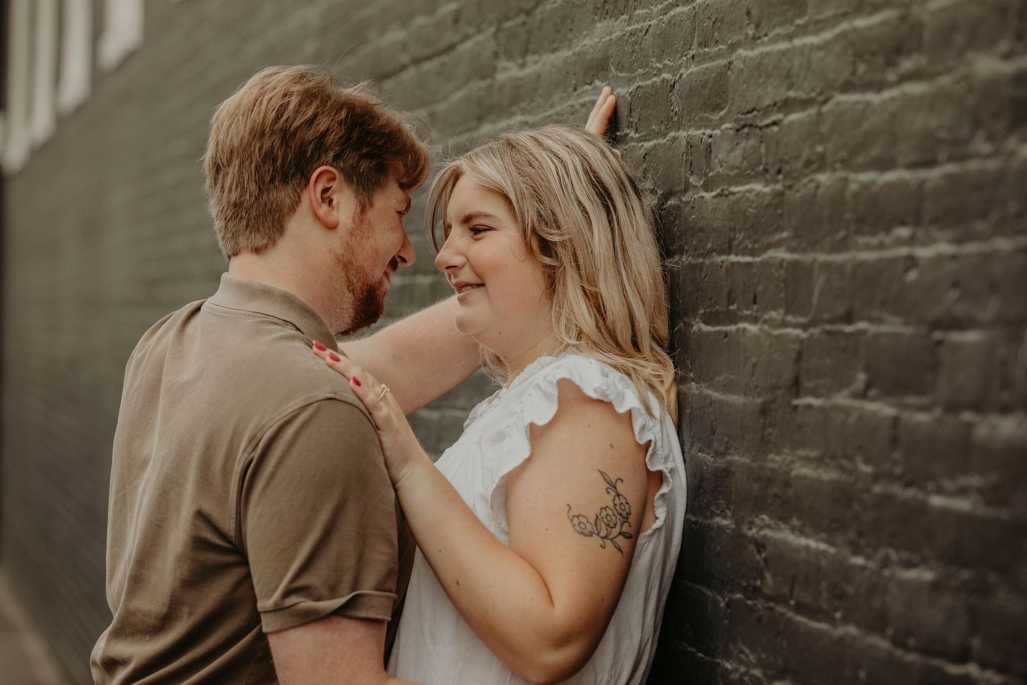 A romantic couple shares an intimate moment against a rustic brick wall backdrop in warm lighting.