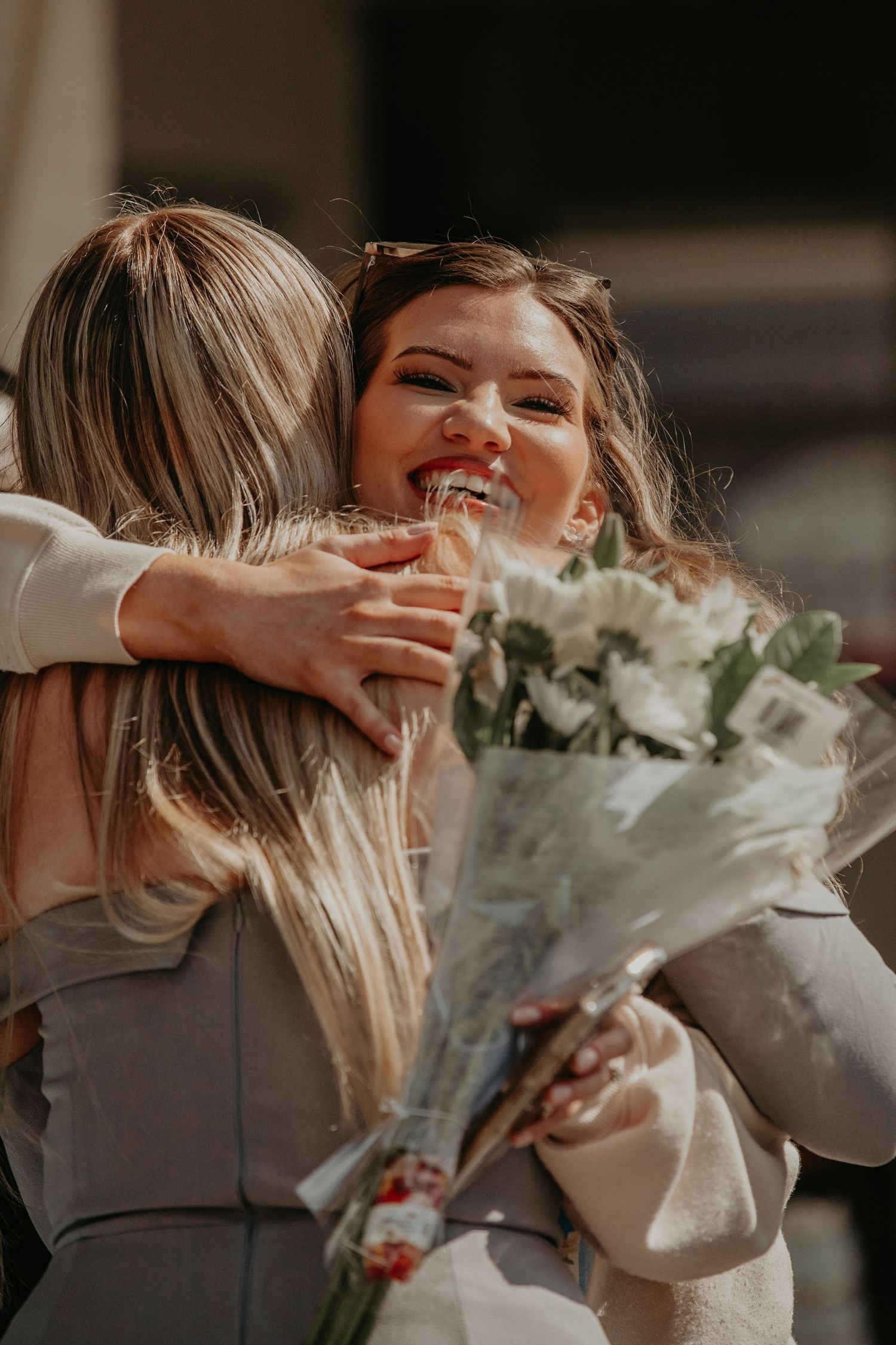 Two friends share a joyful embrace while holding a bouquet of white flowers and eucalyptus on a sunny day.