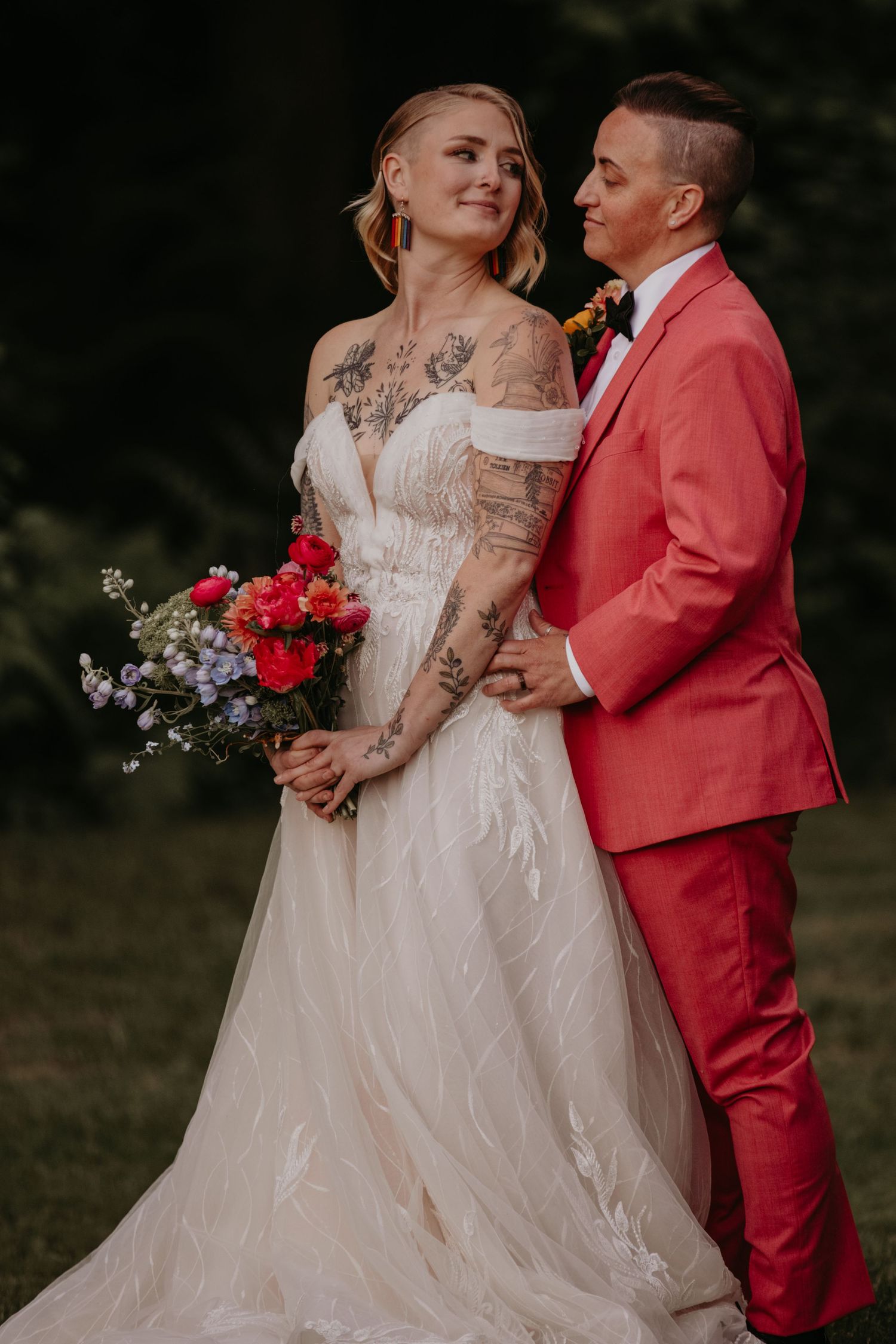 A couple in wedding attire - bride in white dress with tattoos and groom in coral suit - pose with red floral bouquet.