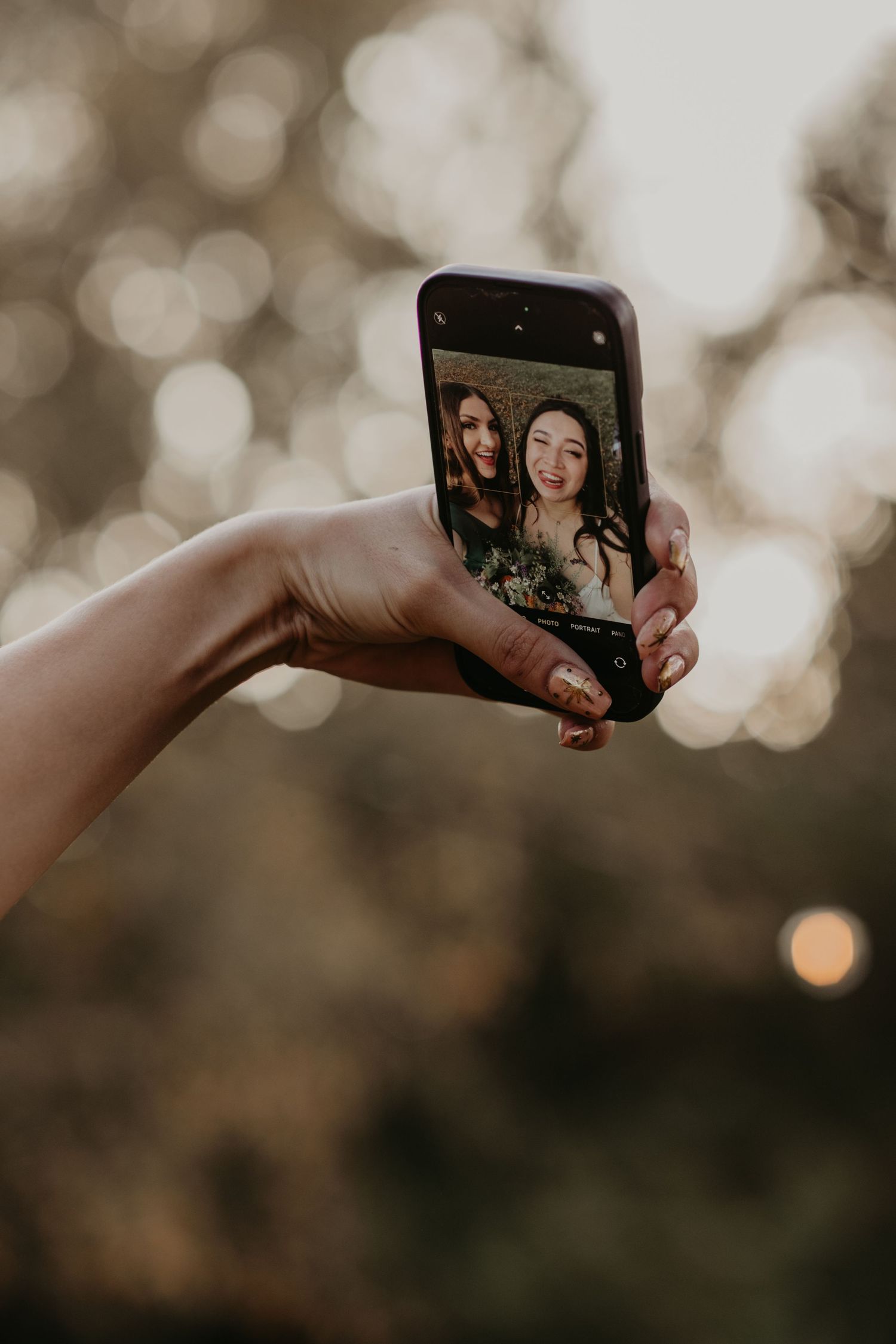 Hand holds up smartphone displaying selfie photo with bokeh lighting effect in background at sunset.