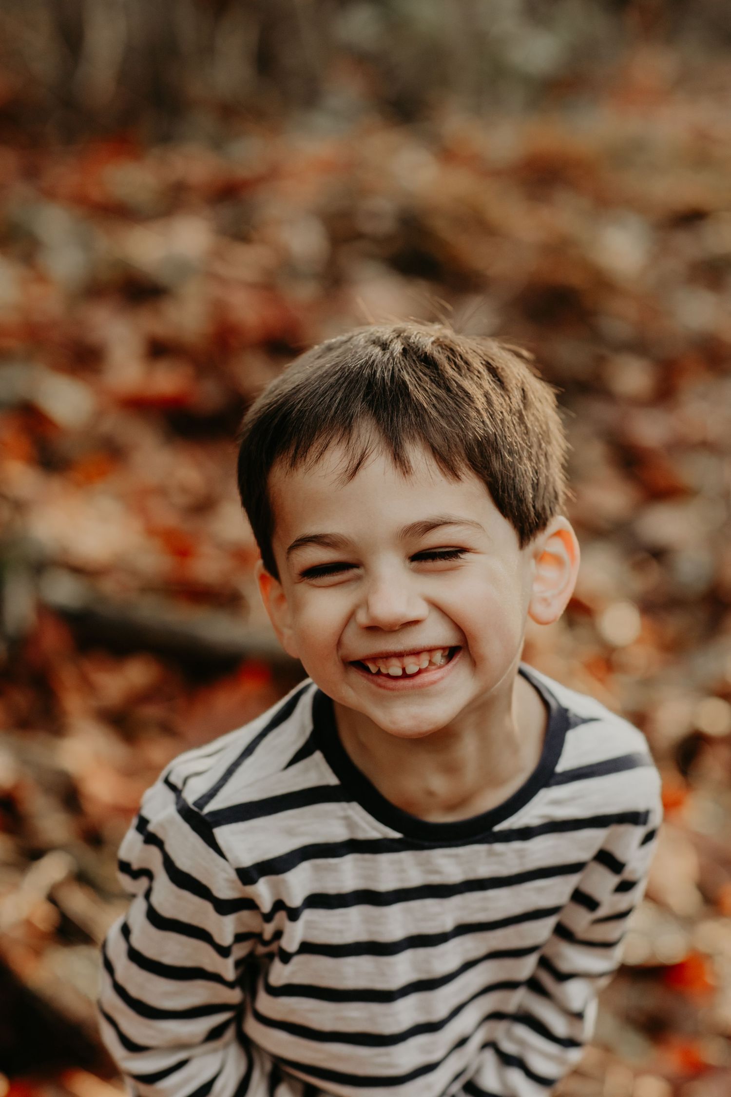 A child in a black and white striped shirt smiles brightly while standing among autumn leaves.