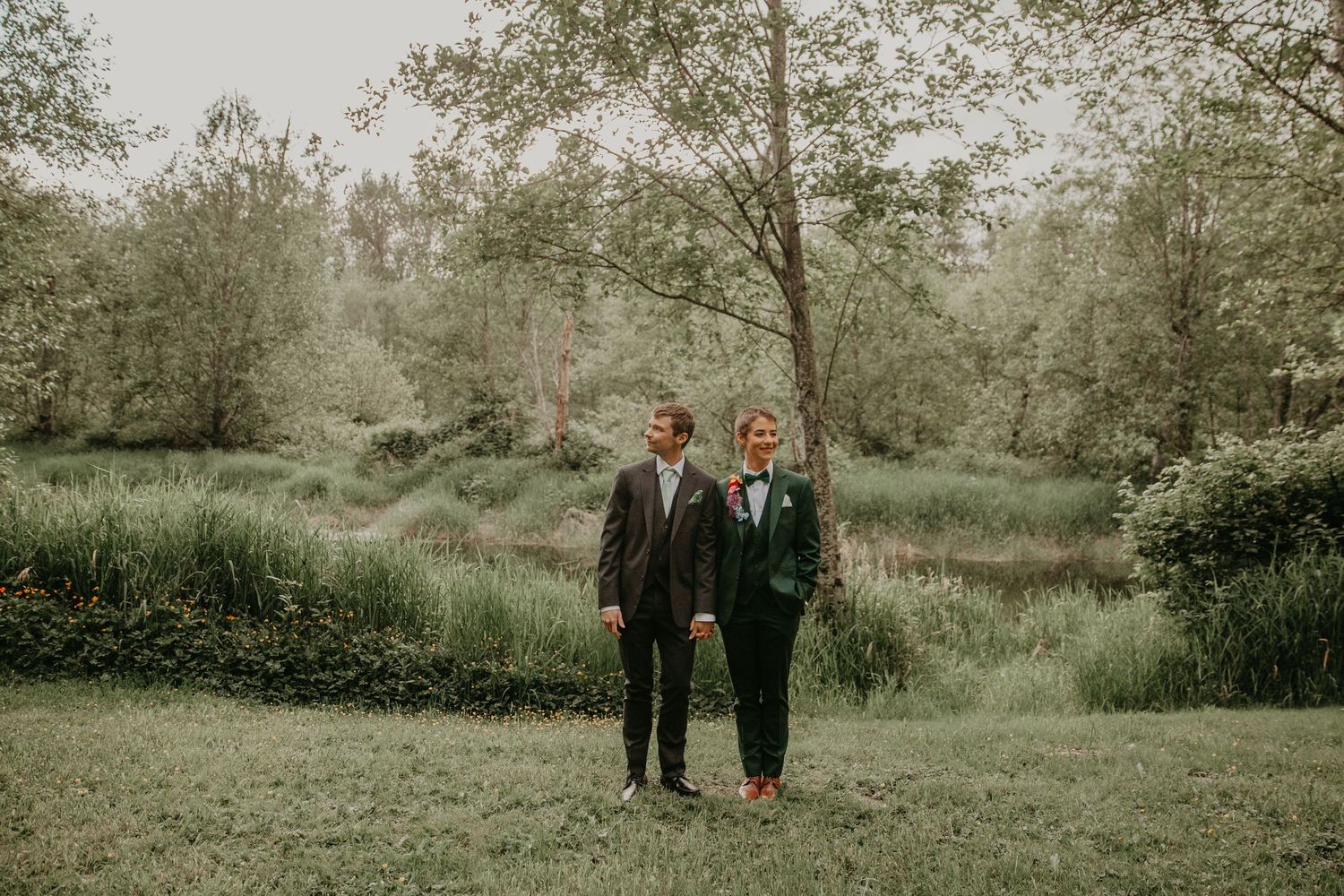 A couple stands together on lush grass in a moody forest setting with muted green tones.