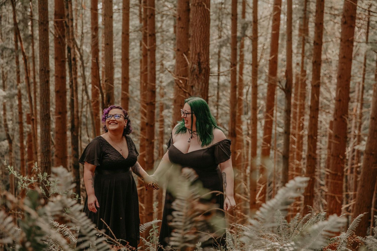 Two people in dark dresses stand laughing together in a forest of tall trees surrounded by ferns.