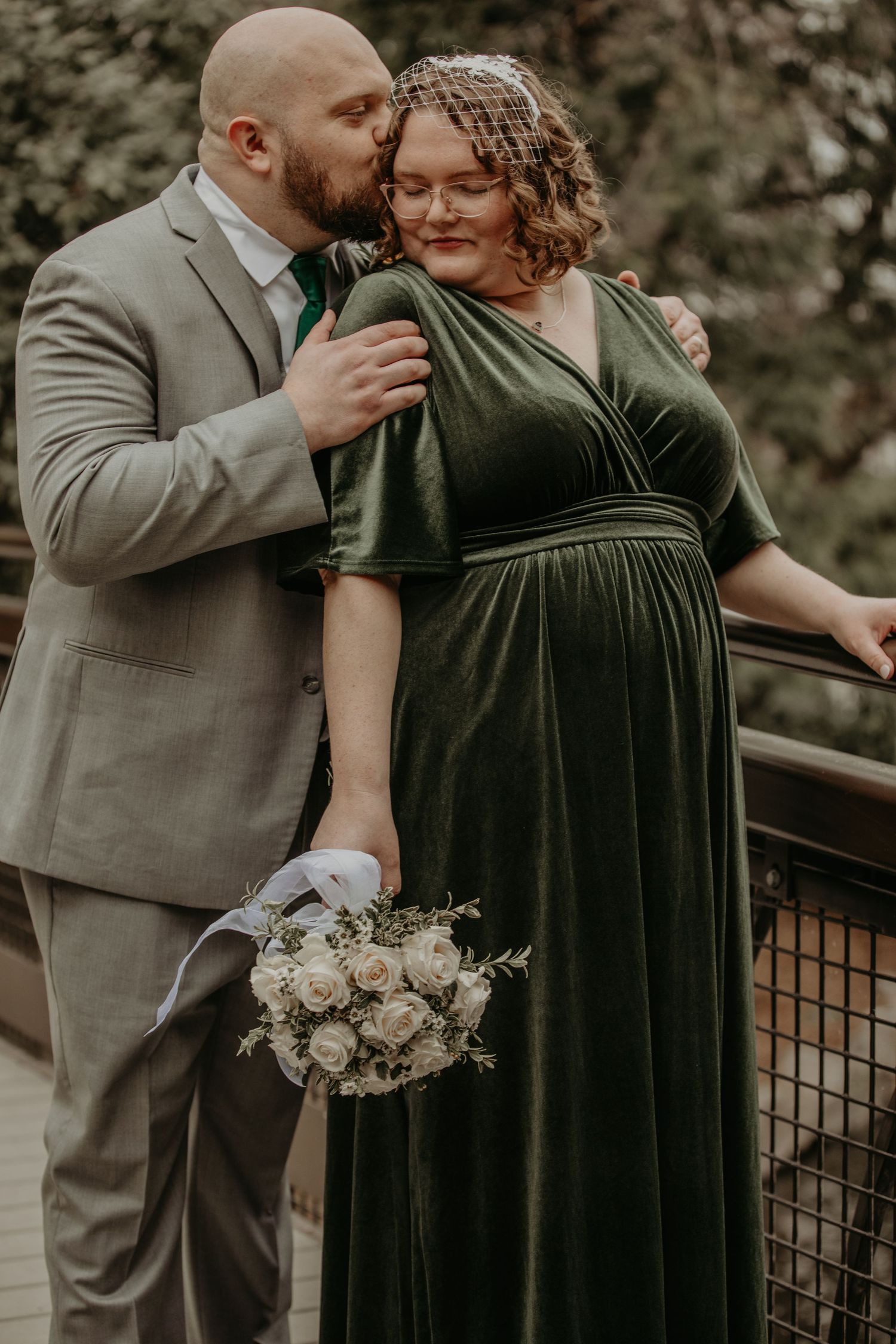 Intimate wedding portrait showing couple in elegant attire with emerald velvet dress and gray suit on wooden deck.