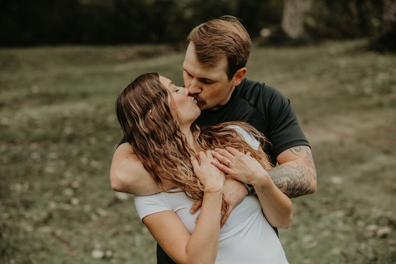 A romantic couple embraces in a tender outdoor setting with soft natural lighting and grassy background.