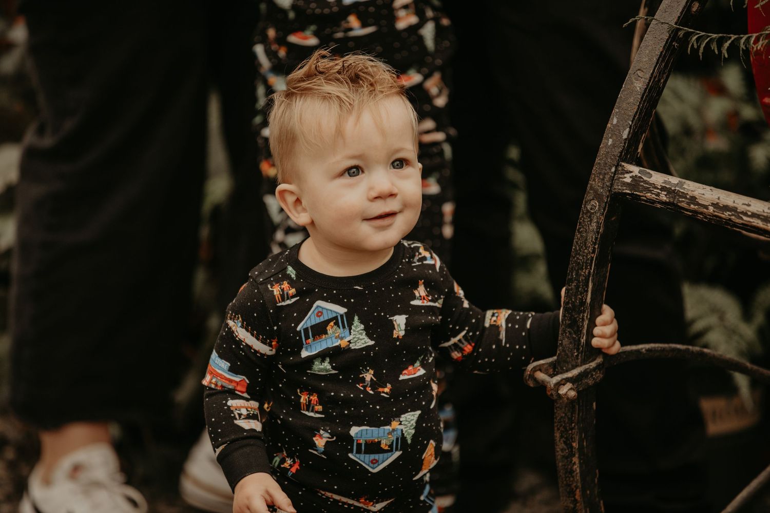 Toddler wearing black sweater with dinosaur print pattern smiles while standing near wooden ladder outdoors.