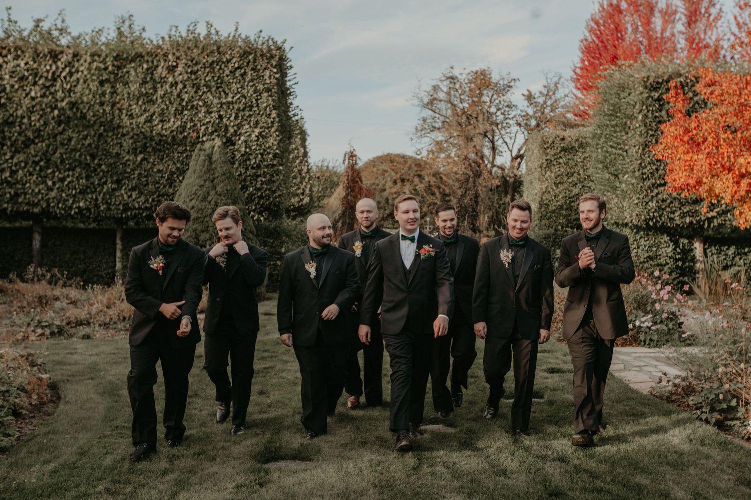 A wedding party in formal black attire walks together through an autumn garden with orange foliage in the background.