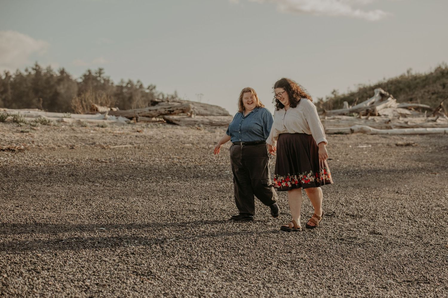 Two people walk together on a rocky beach with evergreen trees and driftwood in the background.