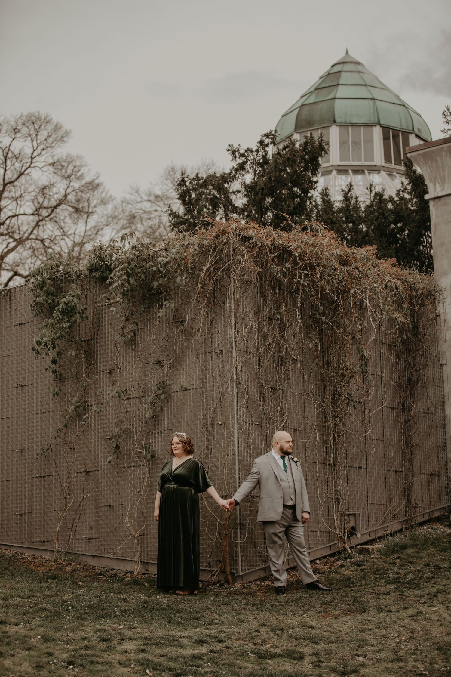 A couple holds hands while standing beside a domed building with climbing vines on the wall.