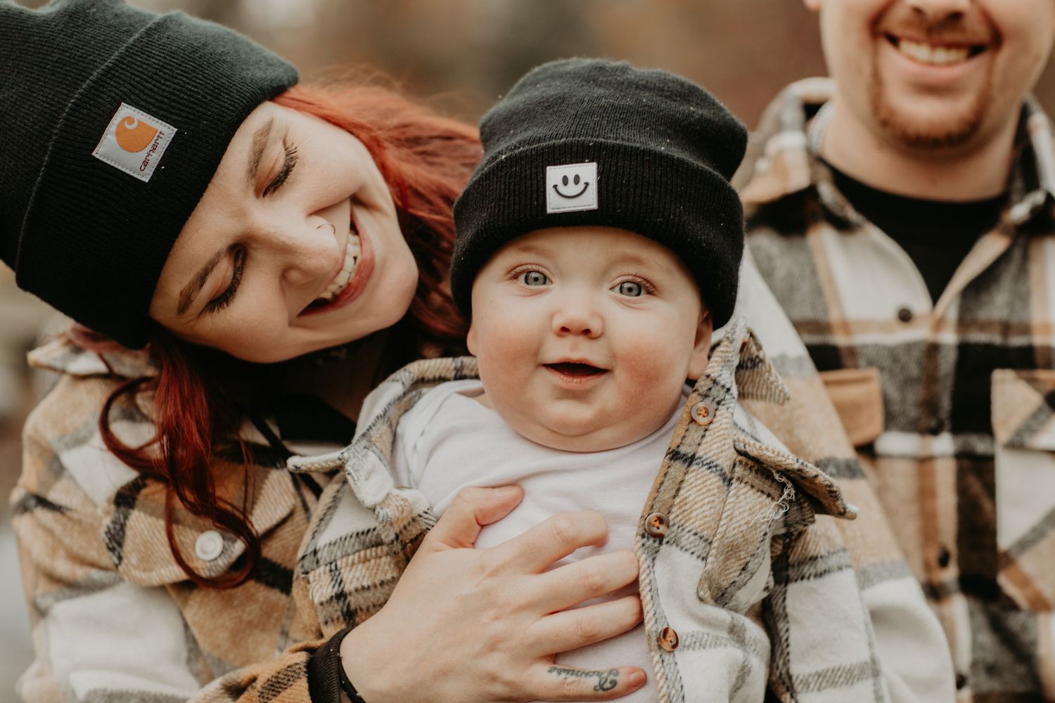 A baby in overalls and a beanie is held lovingly in a candid outdoor family photo.