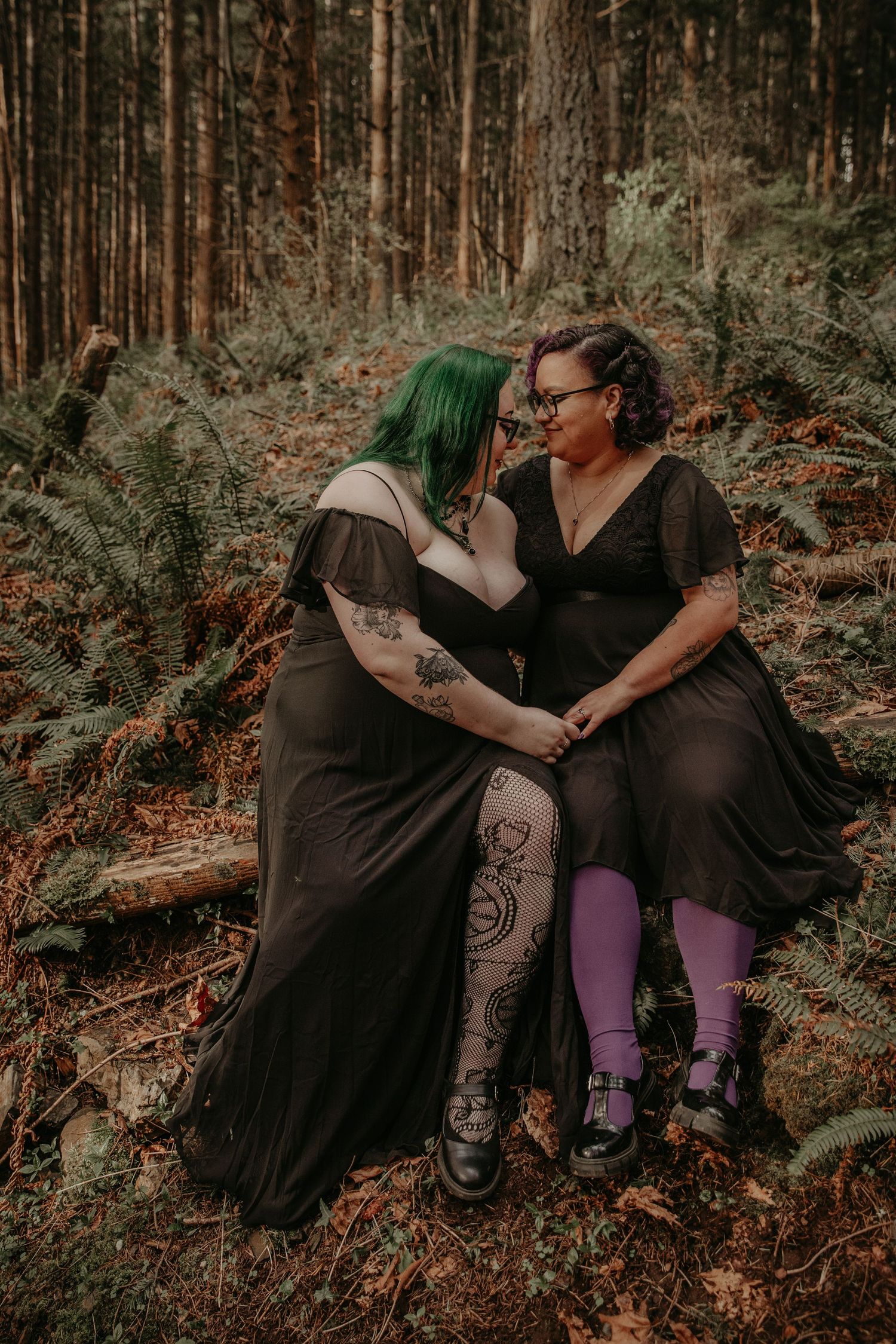 Two people in black dresses sit together in a moody forest setting with ferns and tall trees in the background.