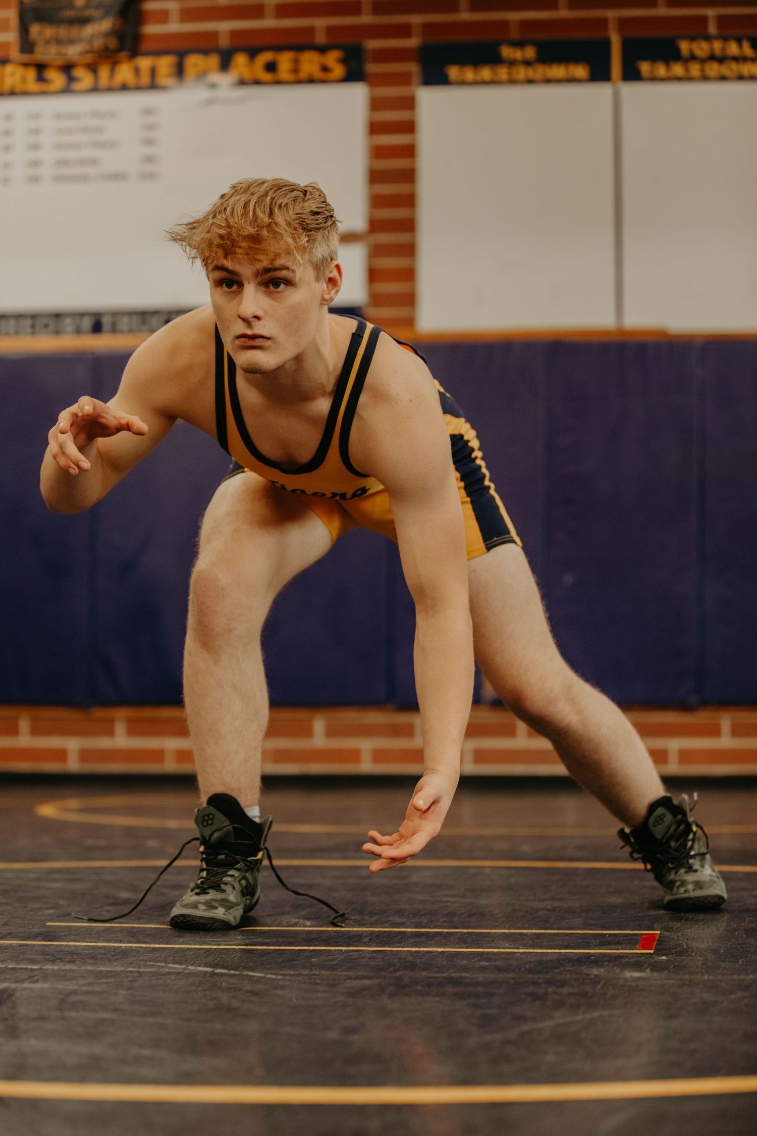 Wrestler in black singlet crouches in ready stance position on purple wrestling mat during match competition.