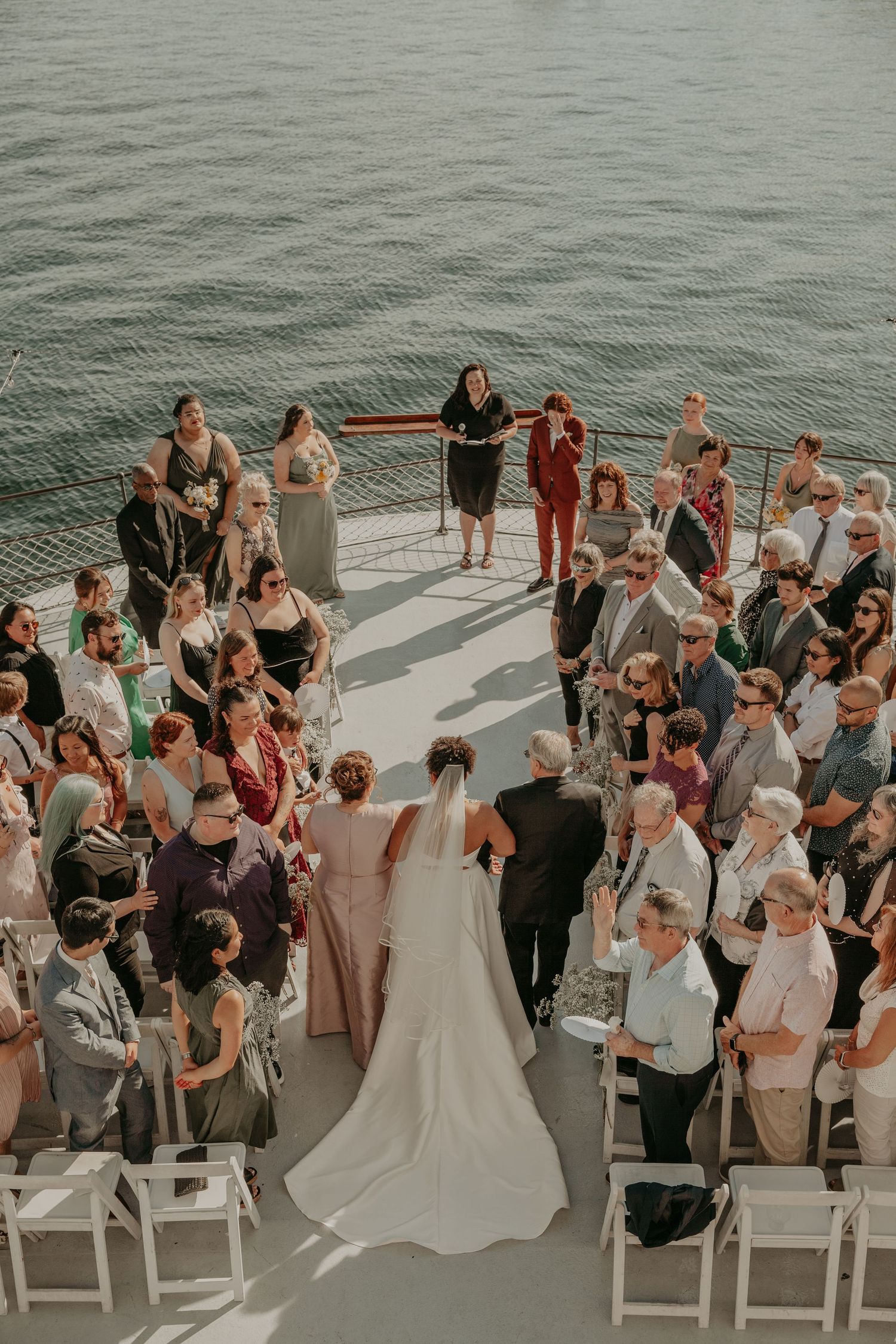 Aerial view of a waterfront wedding ceremony on a boat deck with guests seated in white chairs.