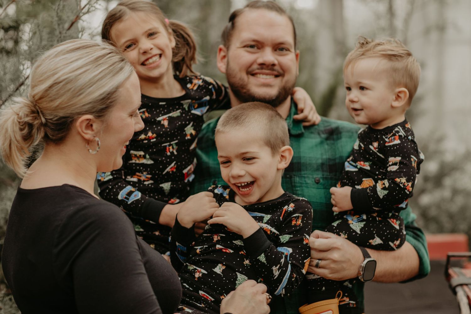 A family shares a joyful moment together outdoors against a wooded background in matching dark patterned clothing.