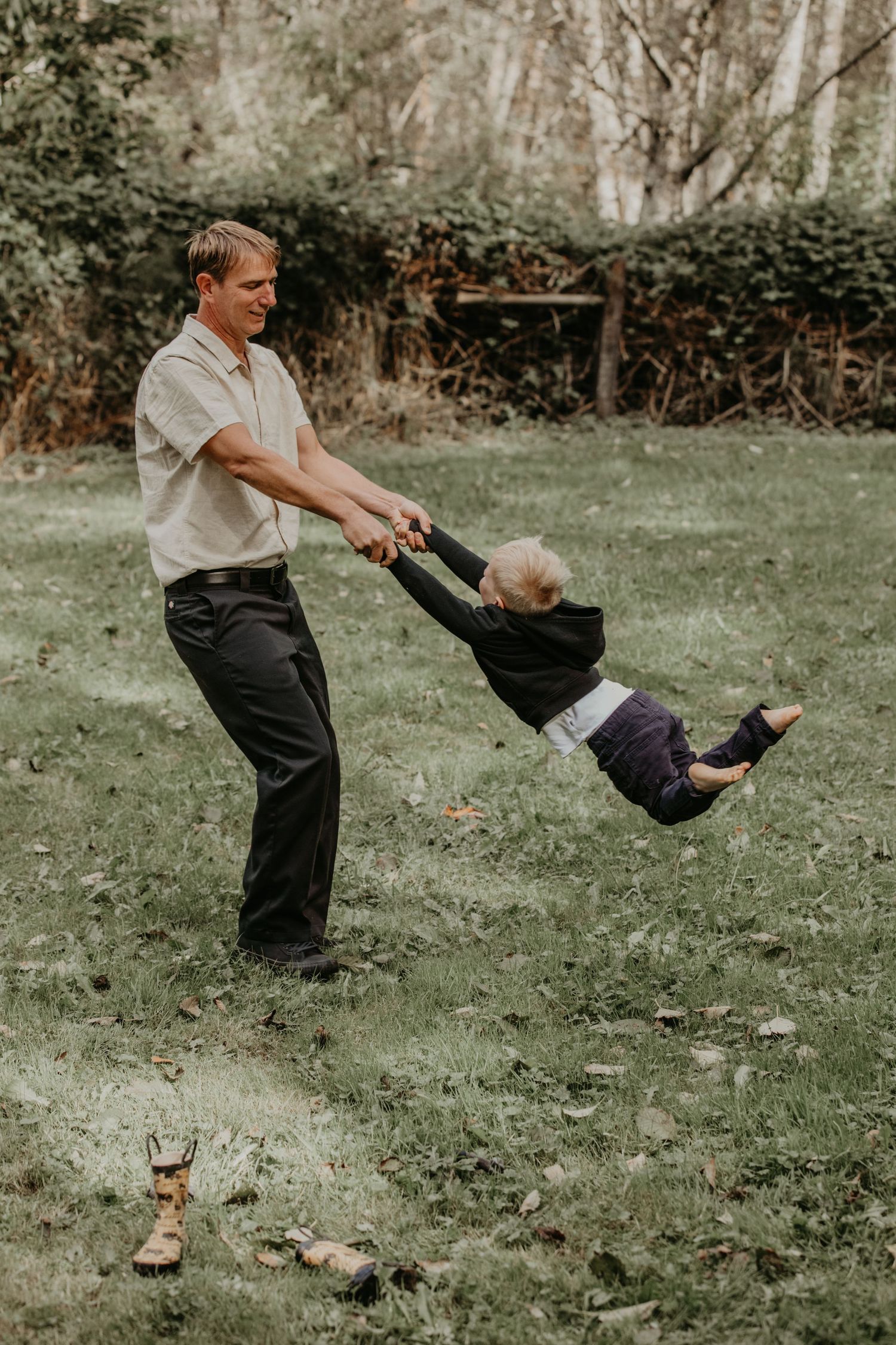A playful outdoor scene shows a guardian swinging a small child by their arms in a green backyard setting.