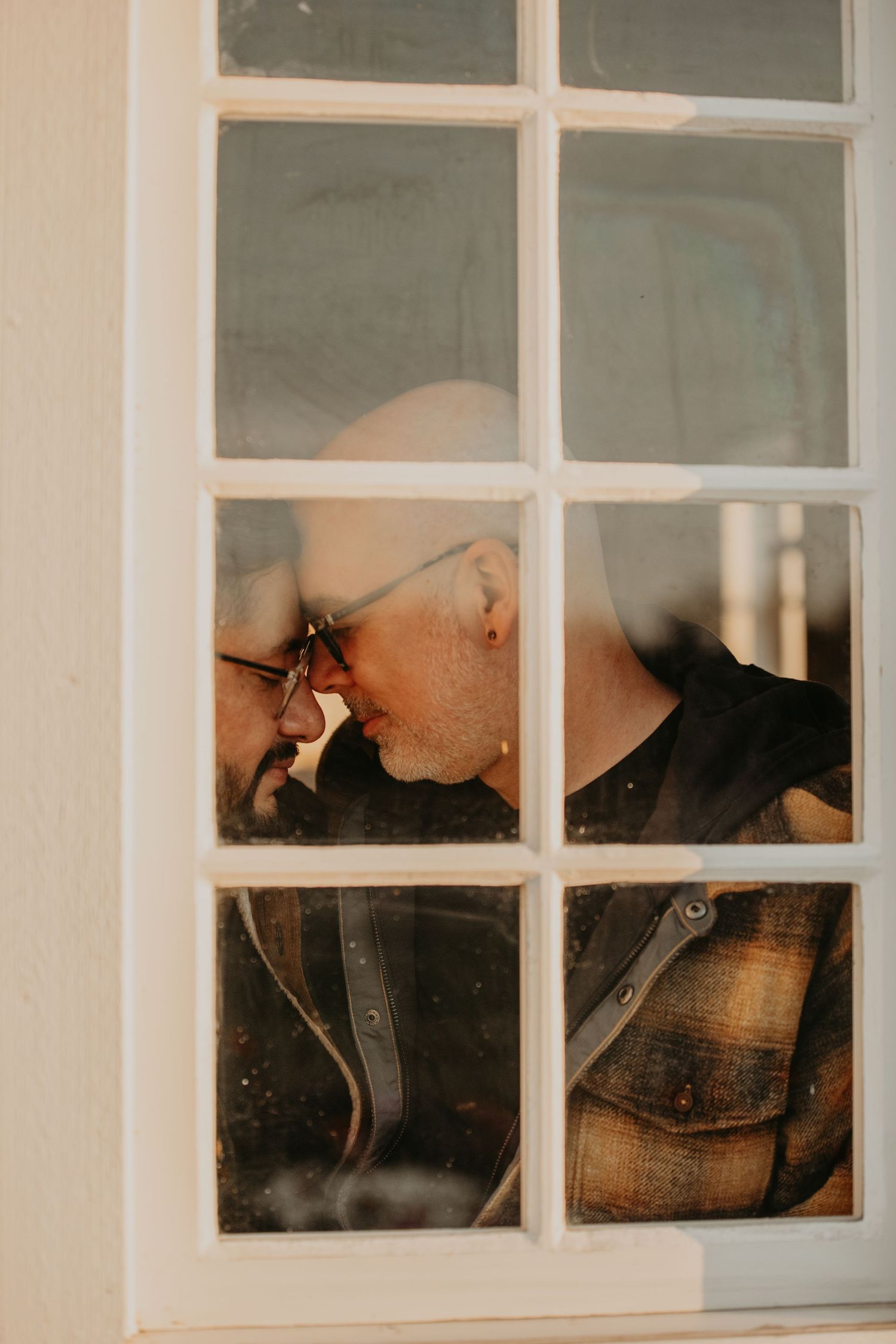 A view through window panes captures an intimate moment between two people in muted tones.