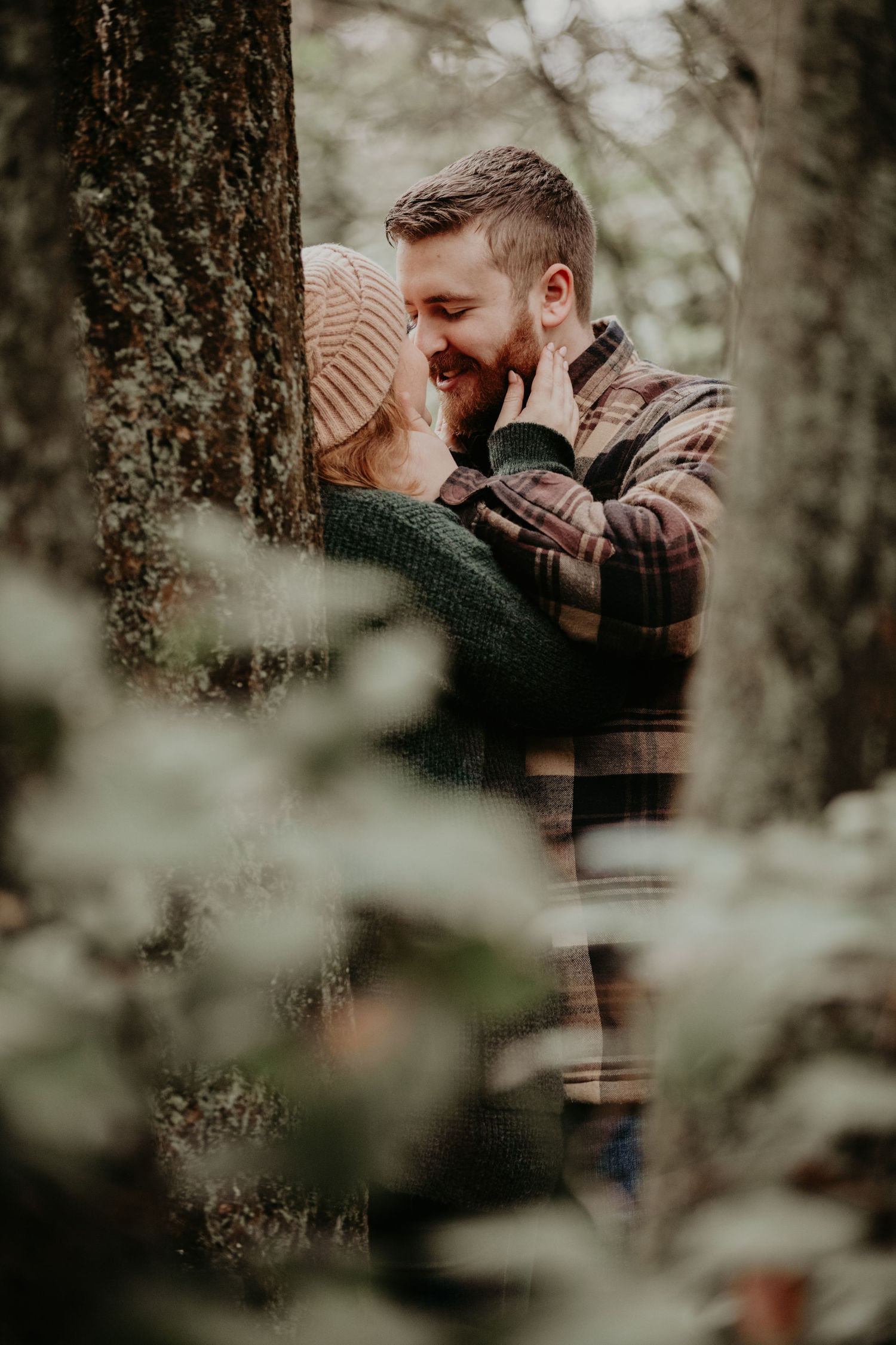 A romantic winter forest scene with a couple embracing beside a tree, wearing cozy sweaters in moody lighting.