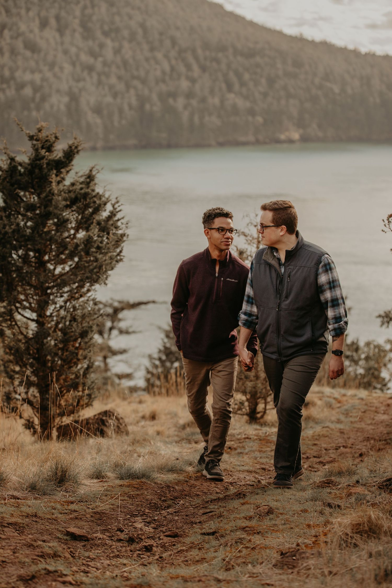 Two people taking a scenic autumn hike along a lakeside trail with mountains and trees in the background.