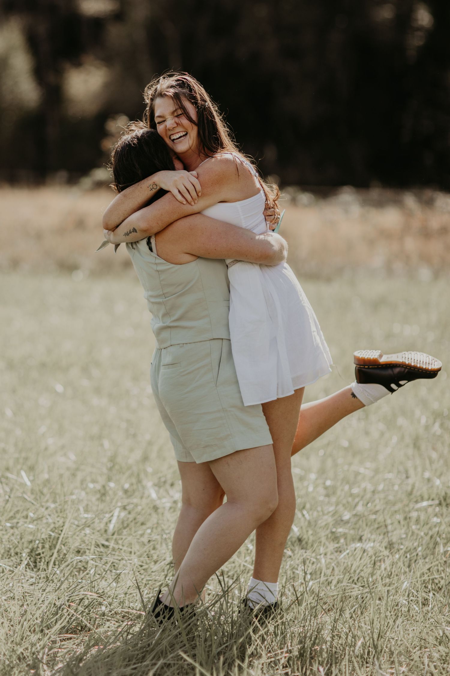 Two people embrace joyfully in a sun-drenched field while wearing casual summer dresses.