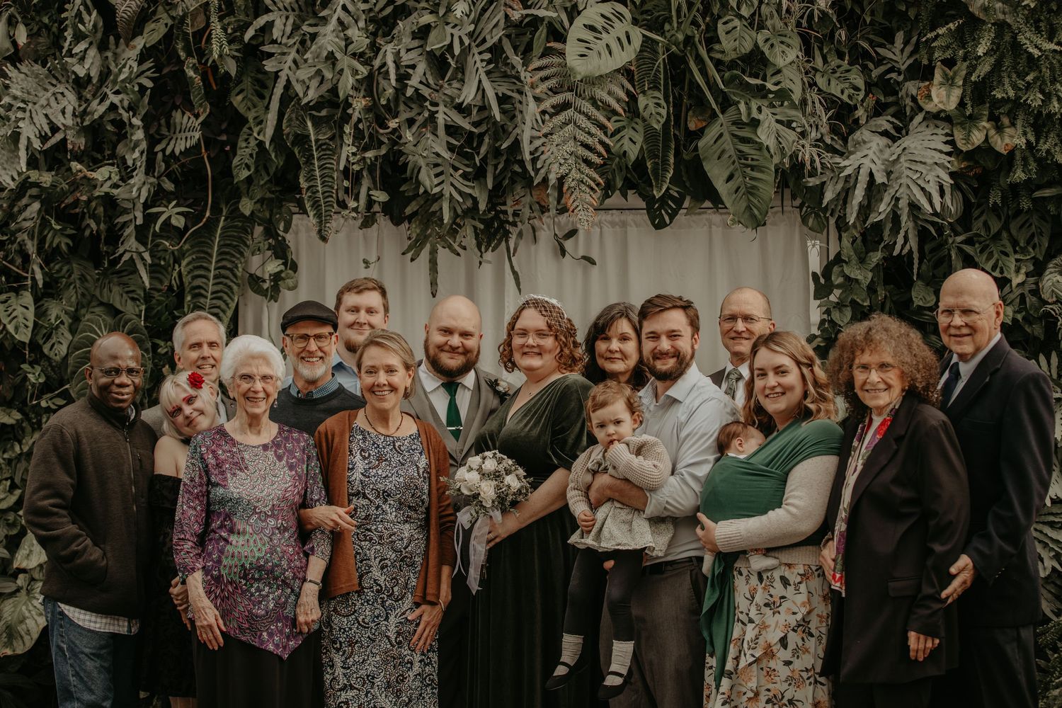 A group of people gathered together outdoors in front of a lush green foliage backdrop at a family celebration.