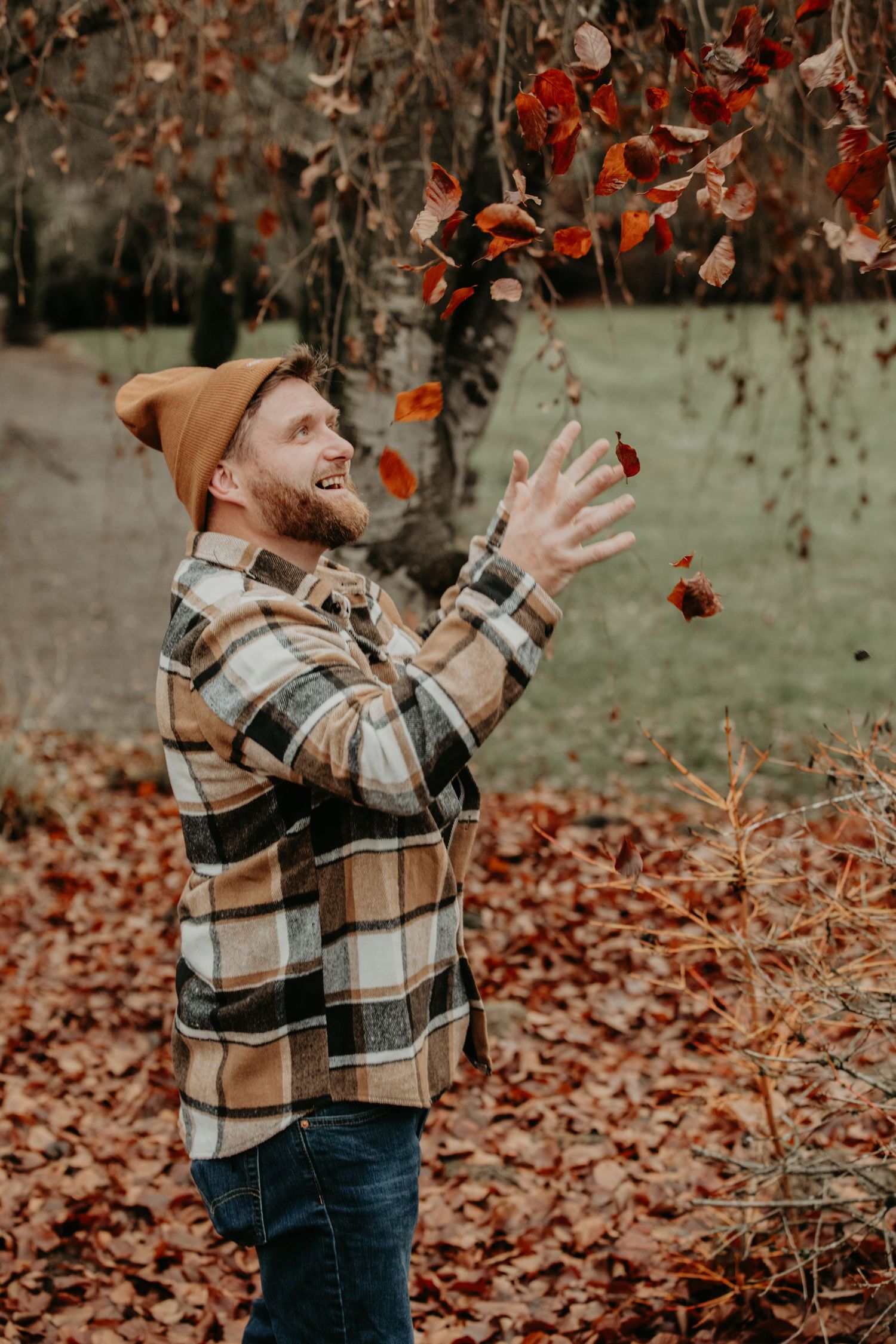 Person in plaid jacket and beanie playfully tosses autumn leaves in the air.