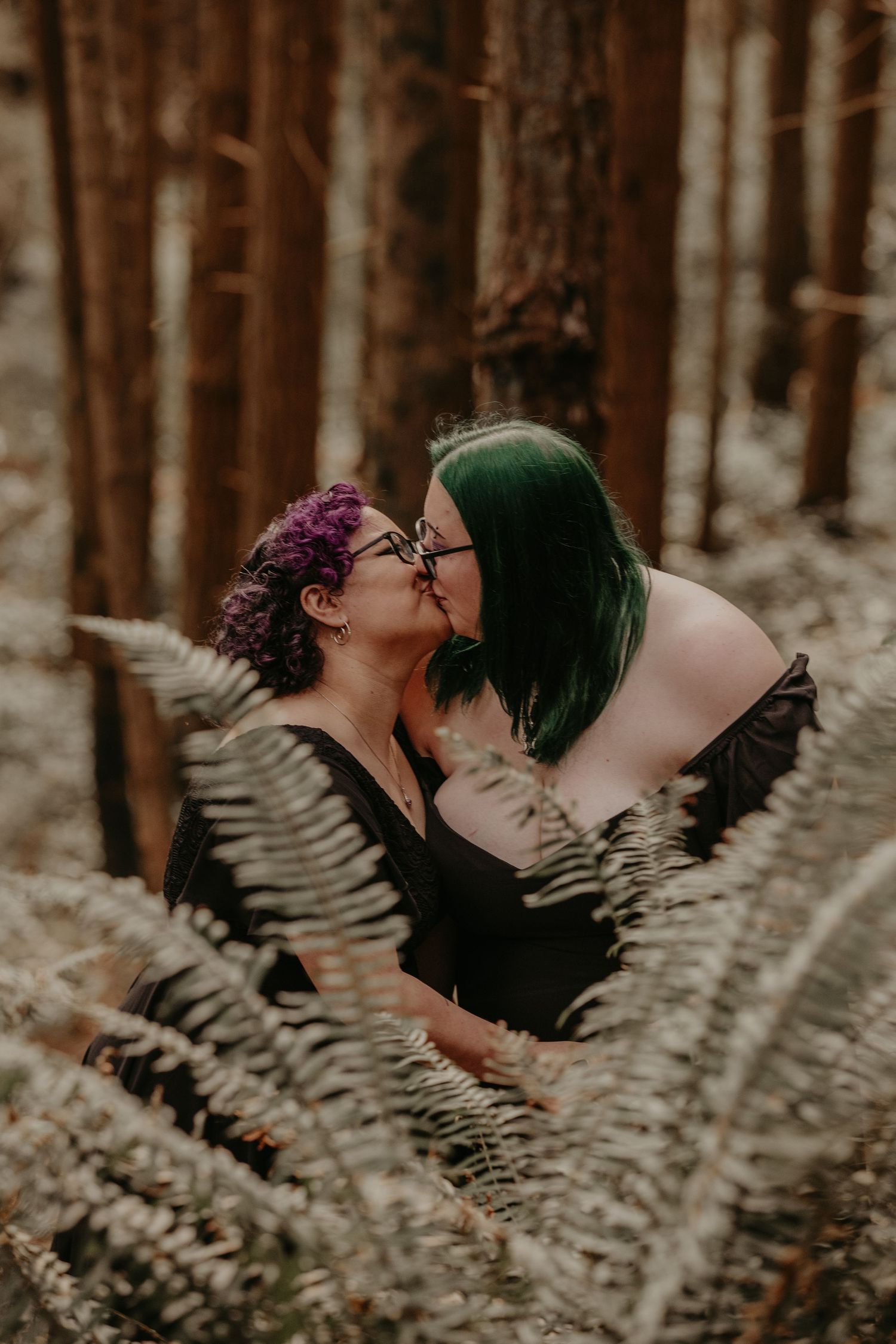 Two people share an intimate moment framed by ferns in a moody forest setting.