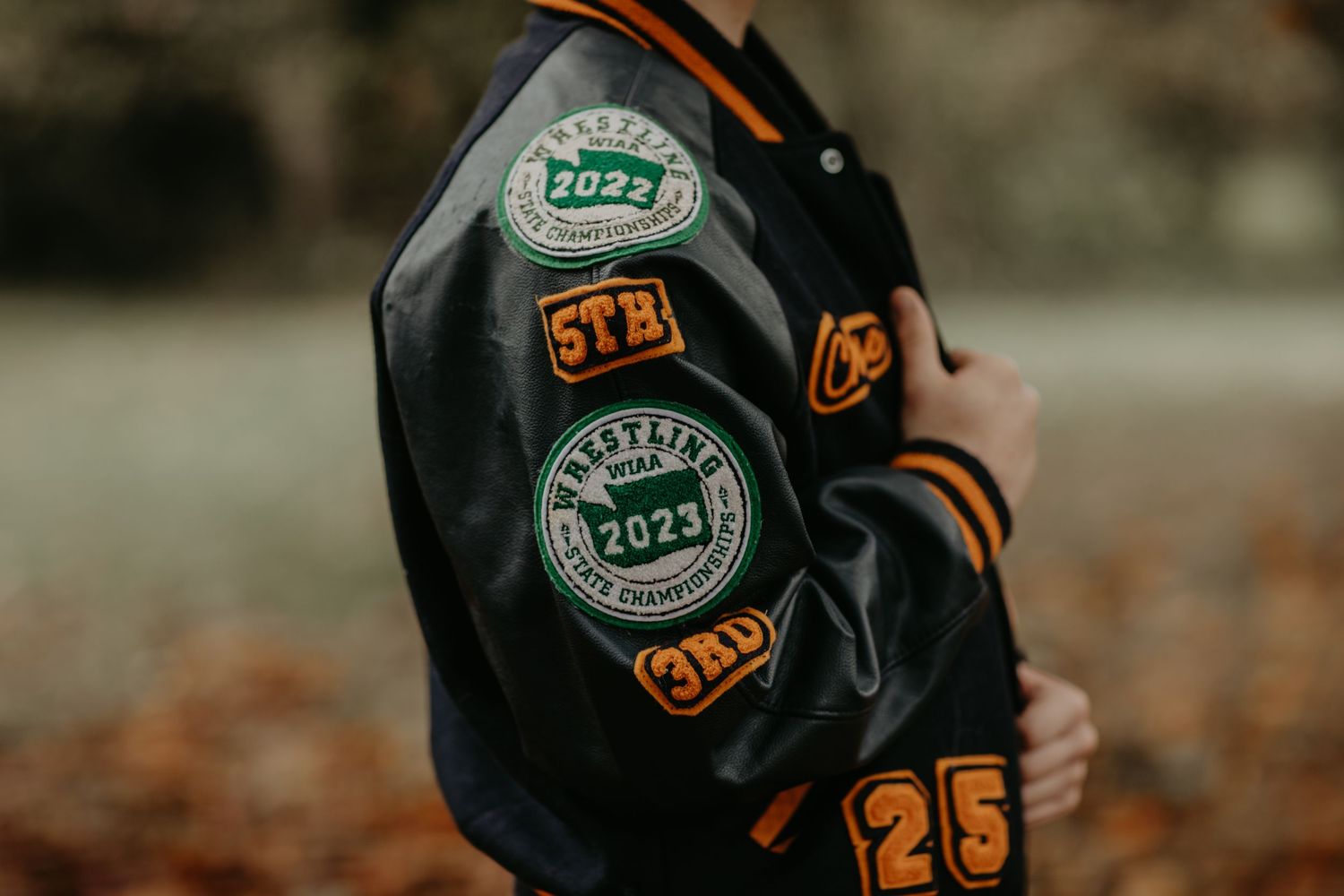 Close-up of a black varsity jacket with orange numbers and green circular patches on the sleeve.