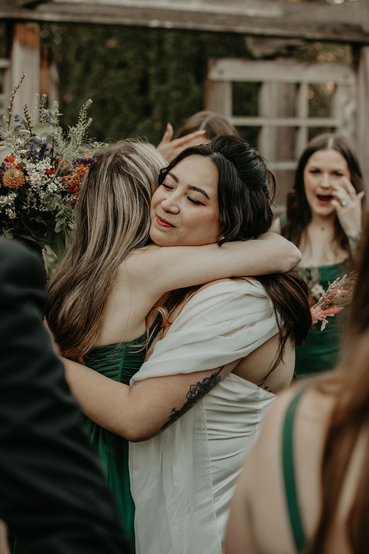 An emotional embrace between wedding attendees wearing white and emerald green dresses at an outdoor celebration.