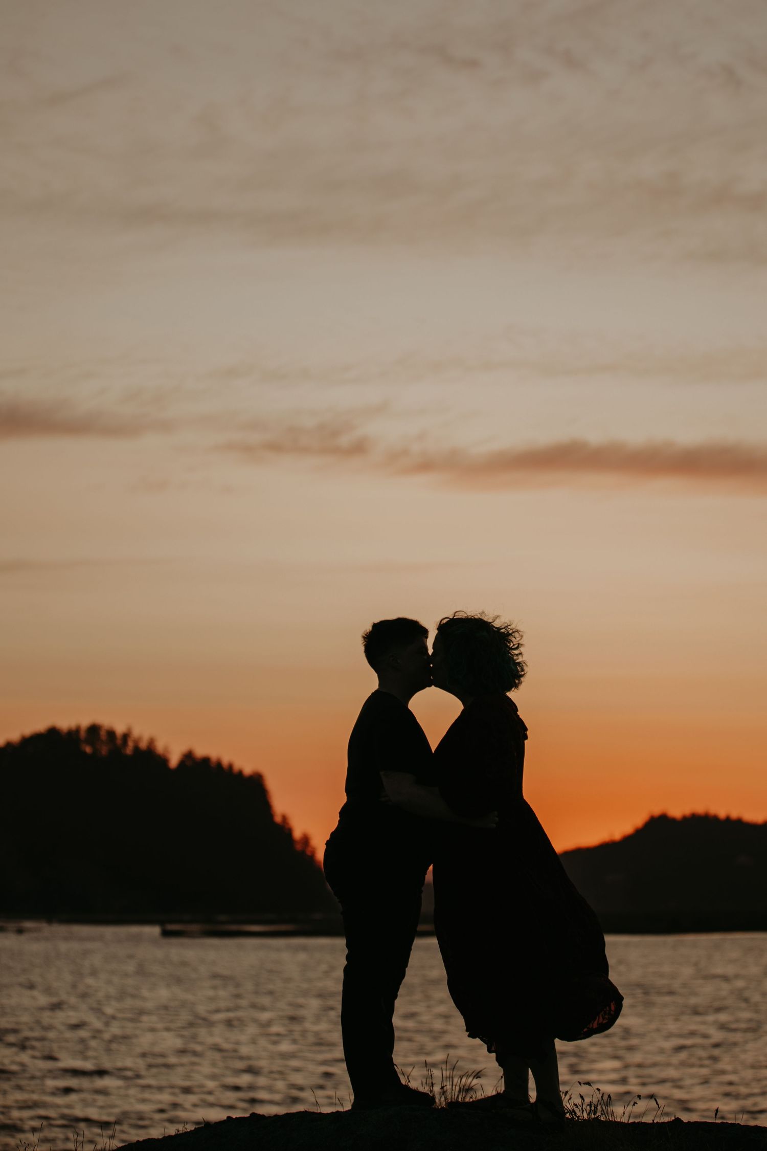 Silhouette of romantic couple standing by water during golden sunset with treeline in background.