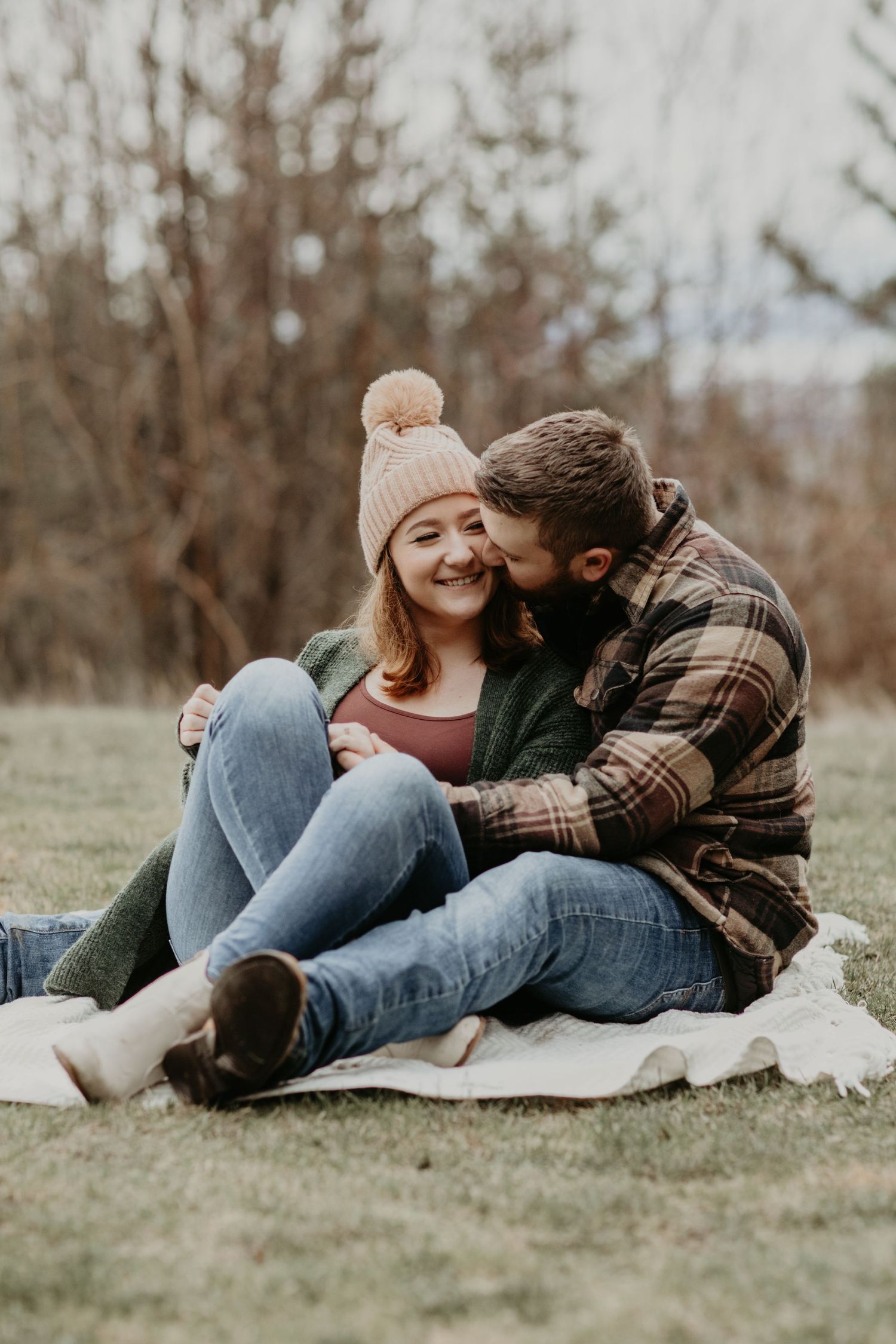 Couple sitting on blanket outside during autumn wearing casual clothes and sharing intimate moment on grassy hill.