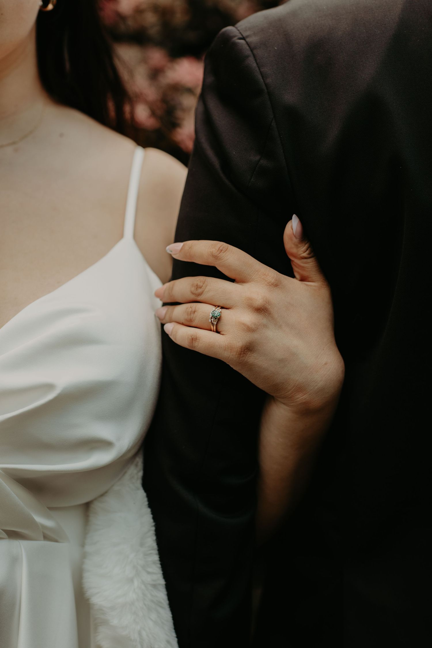 Close-up shot of a couple embracing, with focus on hands and wedding ring against white dress and black suit.