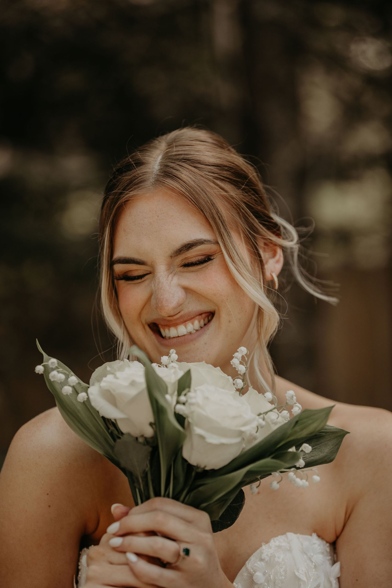 Joyful bride in white strapless dress laughs while holding elegant bouquet of white roses and lily of the valley.