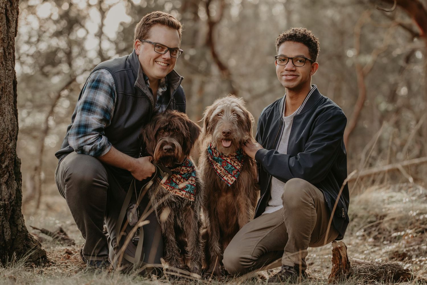 Two dogs with bandanas pose with their owners in outdoor fall setting with brown and sepia tones.