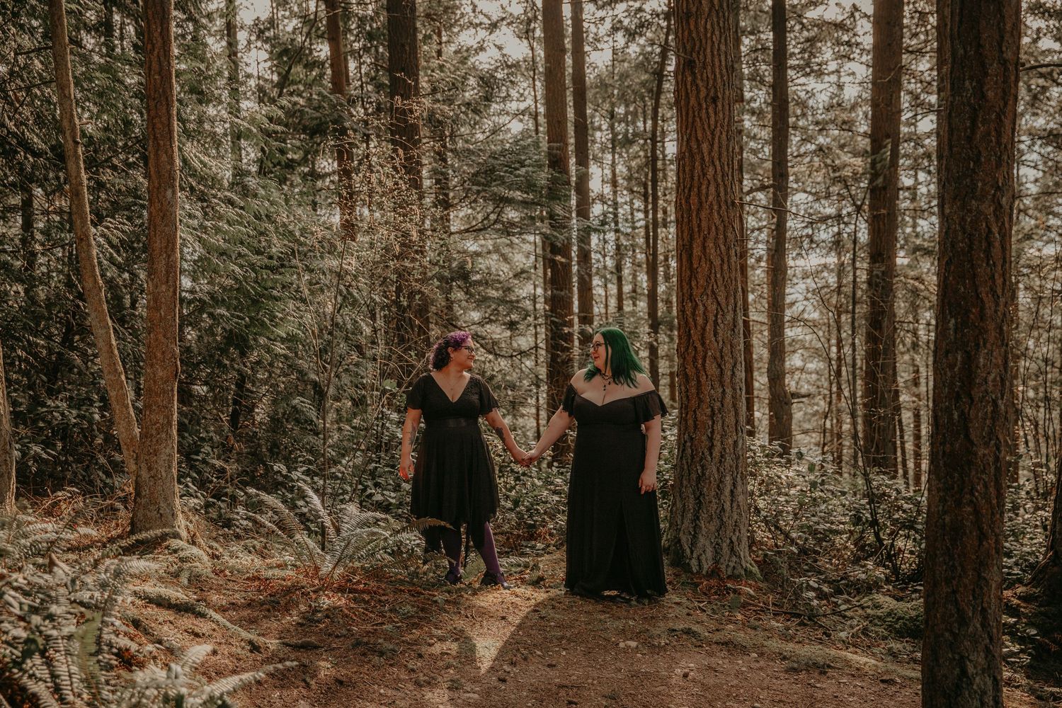Two people in black dresses explore a serene forest trail surrounded by tall evergreen trees at sunset.